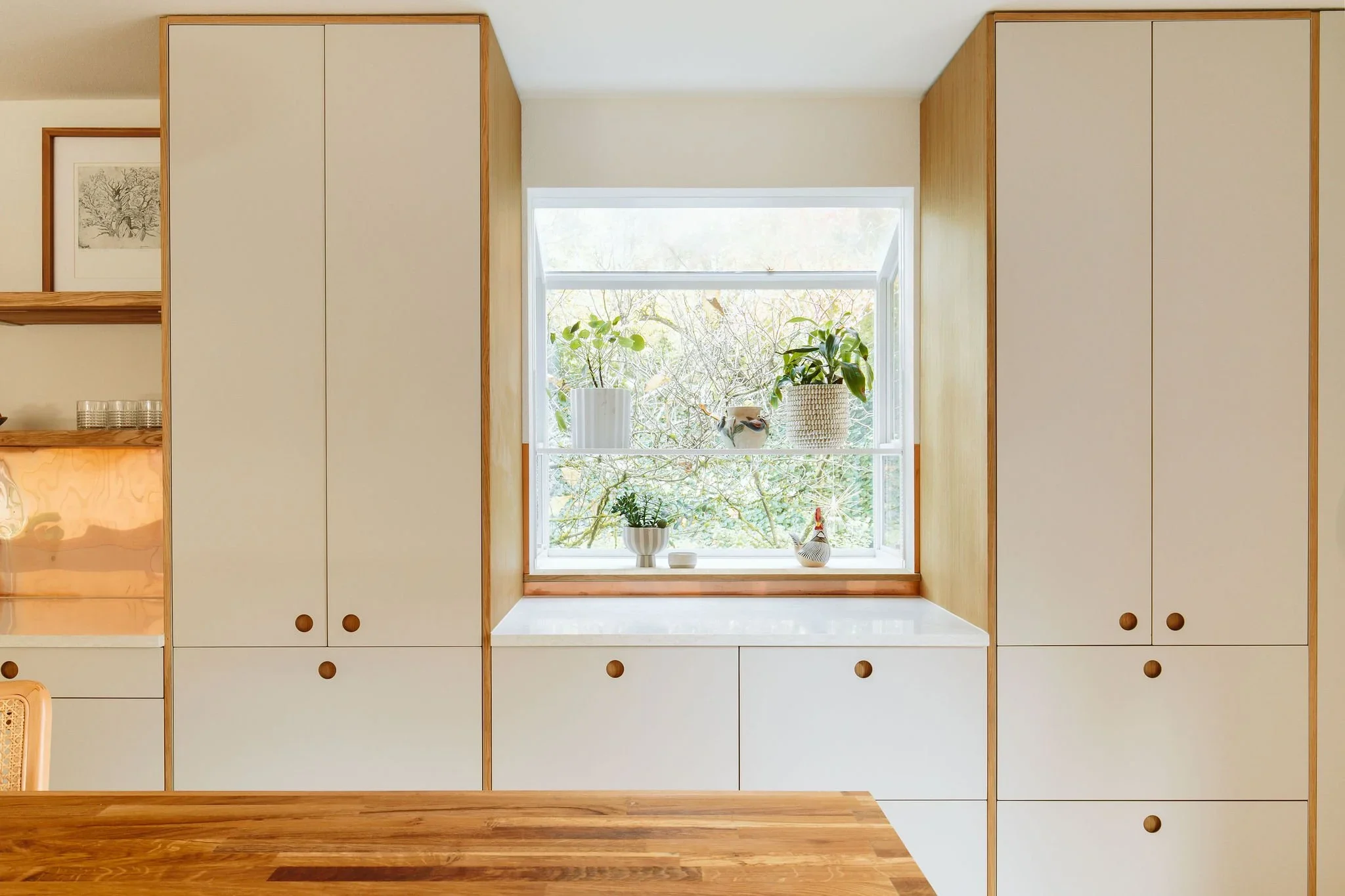 A kitchen with white cabinets, wooden knobs, a window with potted plants, and a wooden countertop in the foreground.