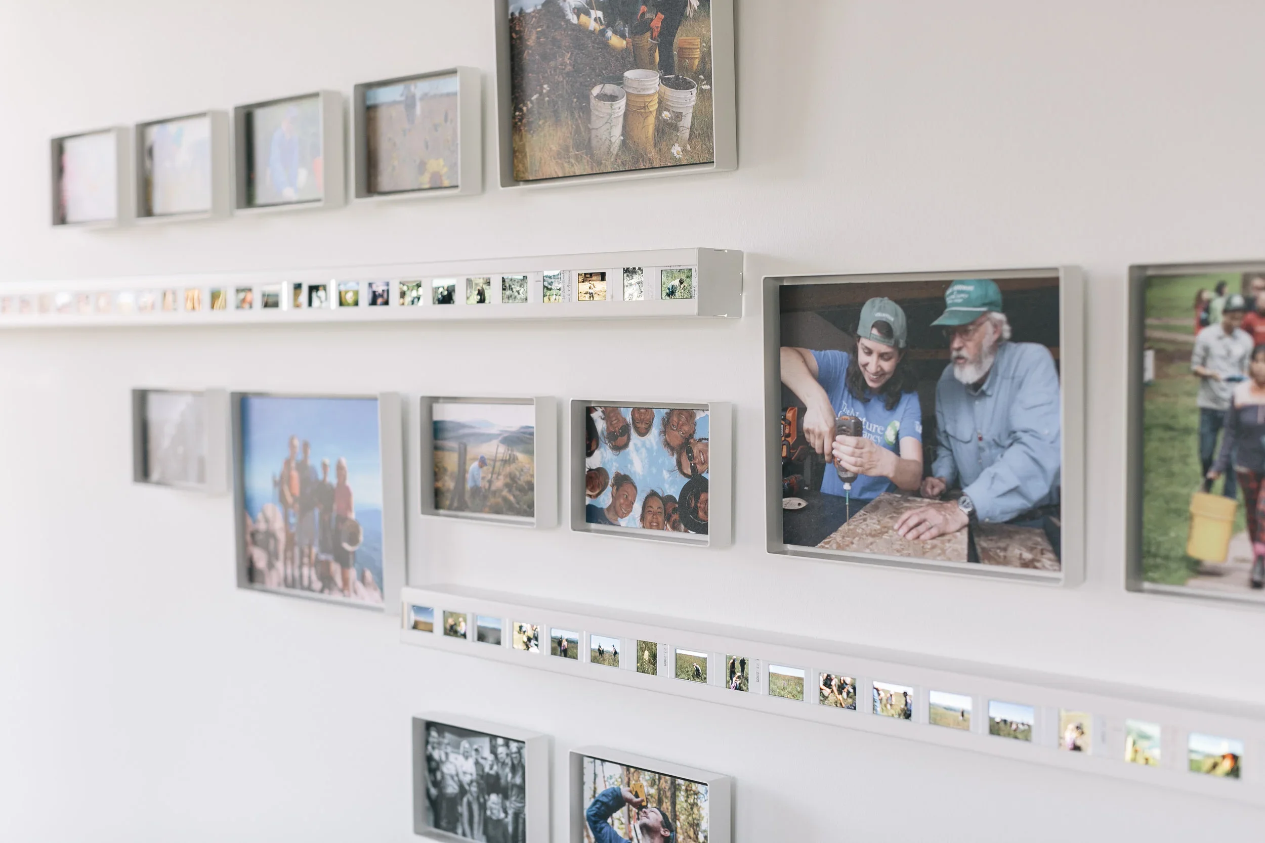 A wall display with multiple framed and unframed photographs, including images of people outdoors and engaging in activities.