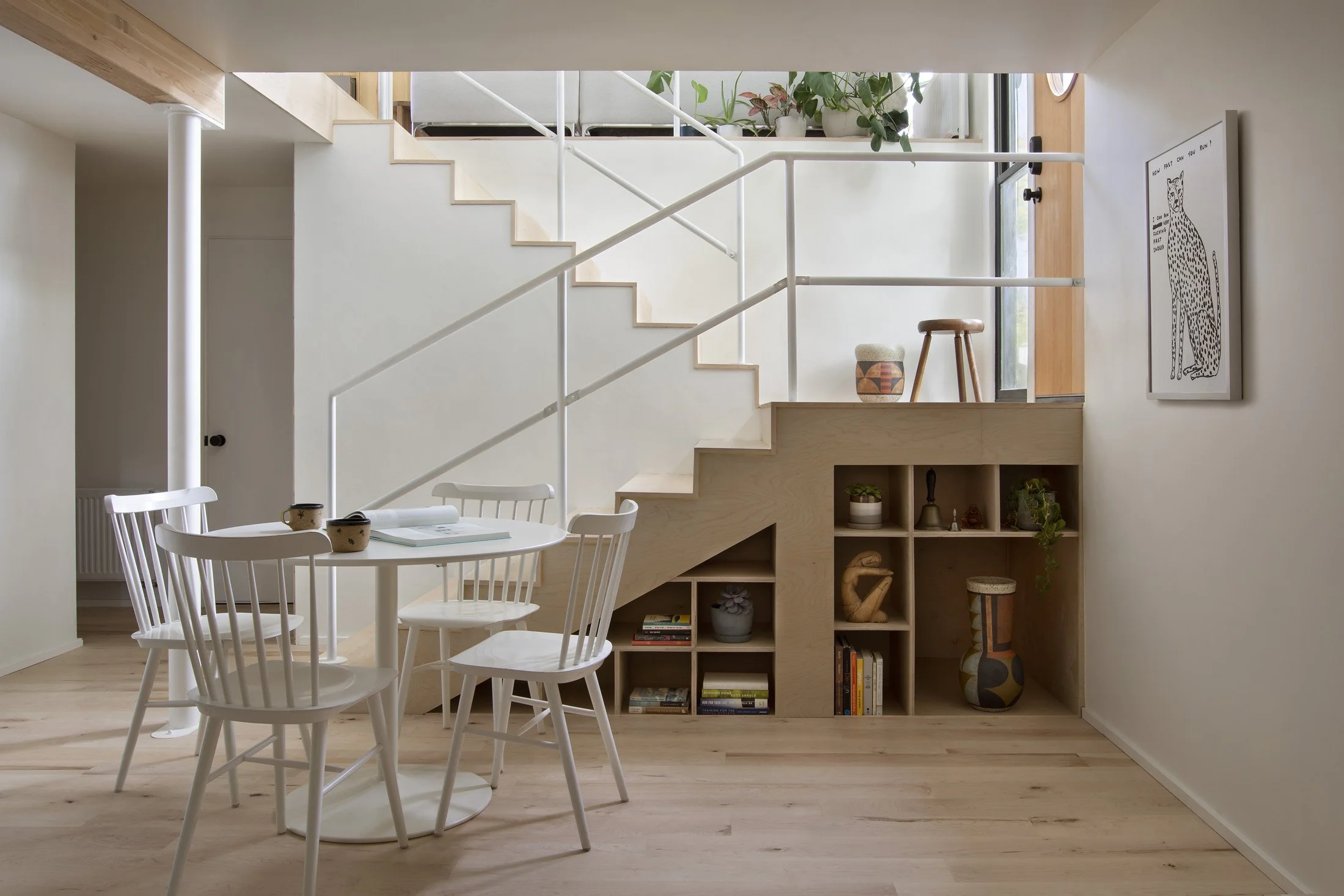 Modern interior with a white round dining table, four white chairs, and built-in wooden stairs with an open shelving area underneath. Decor includes plants, artwork, and books.
