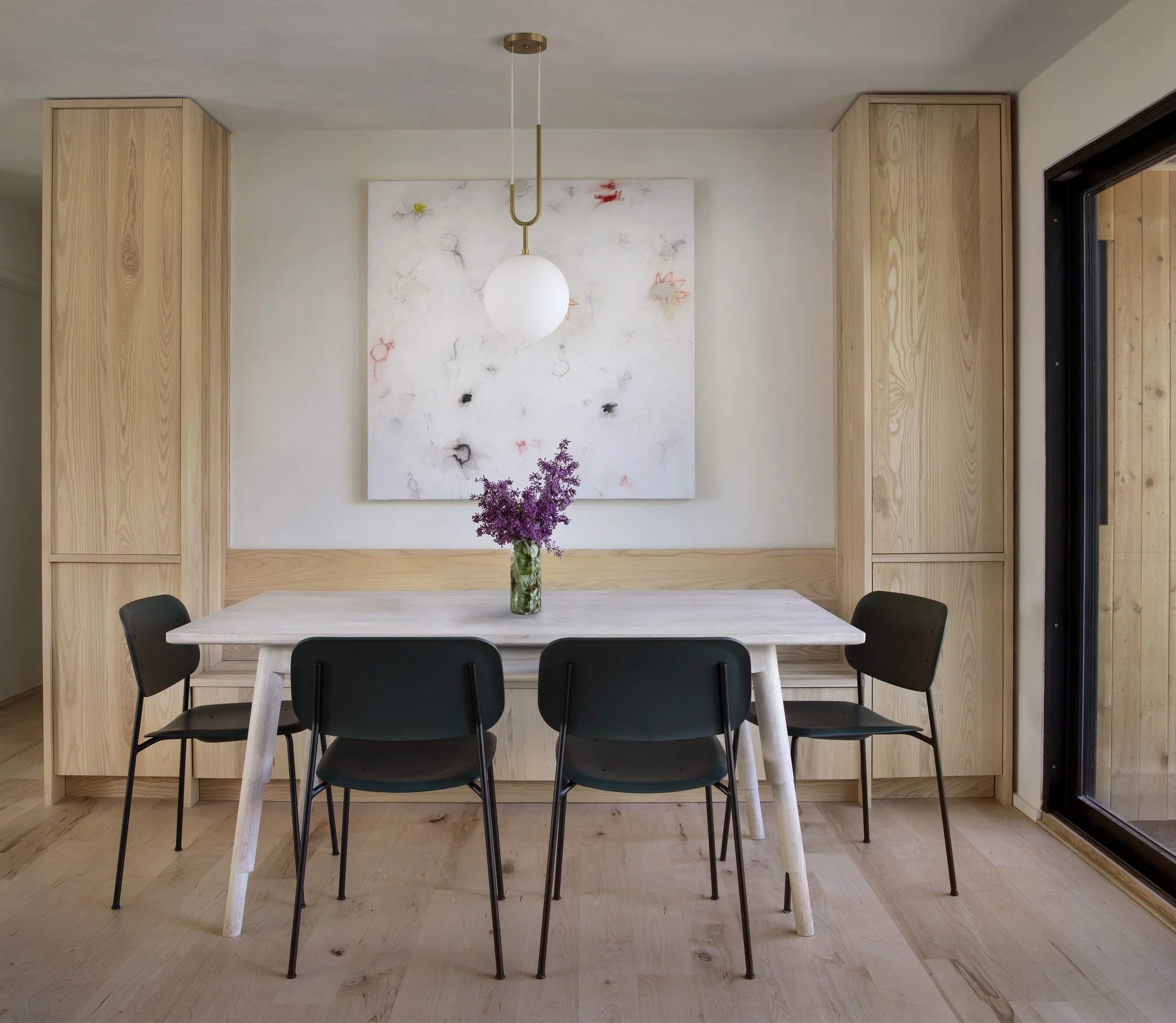 Minimalist dining room with a white wooden table, six black chairs, a glass vase with purple flowers, built-in wooden cabinets on both sides, and a large abstract painting with colorful marks on the wall behind. A white globe pendant light hangs from the ceiling. Sliding glass door to the right.