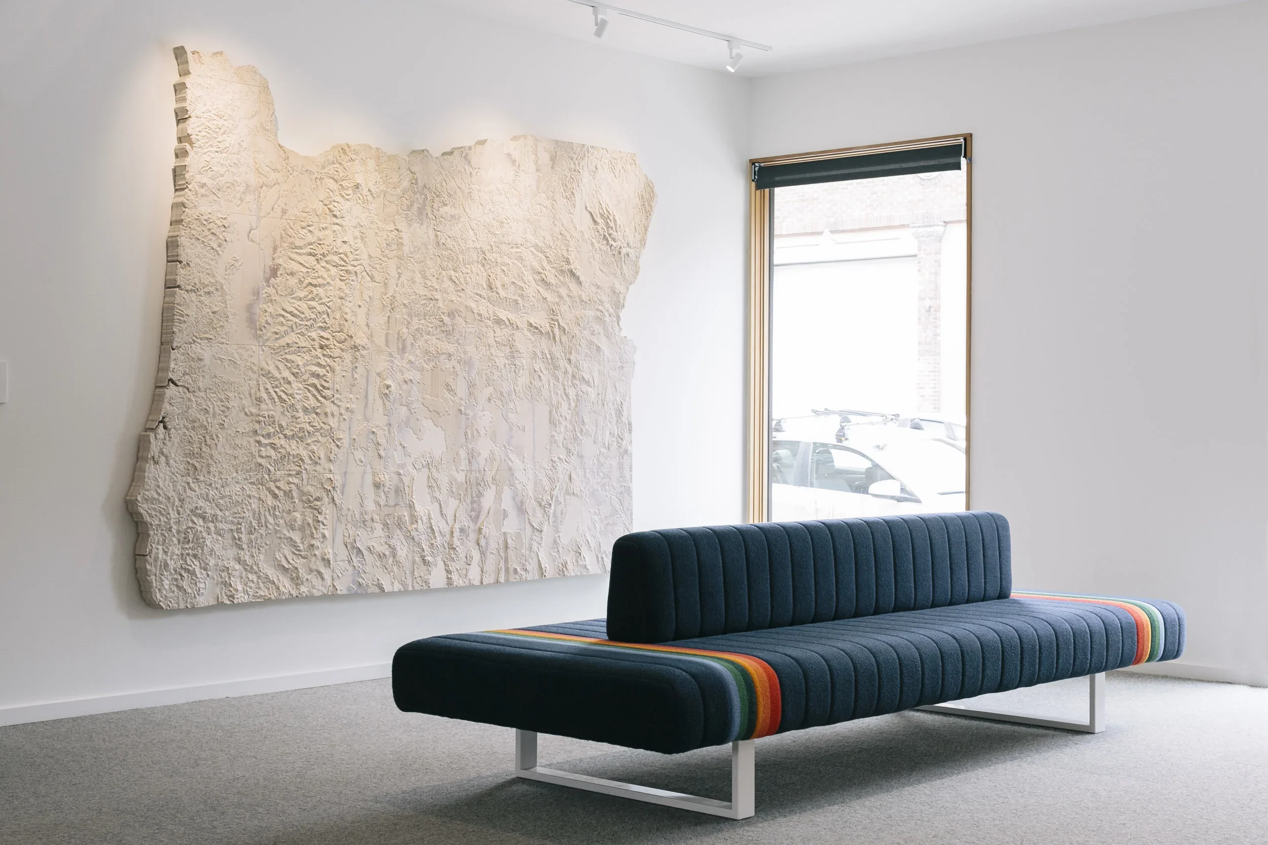 A modern interior with a textured abstract wall art, a blue upholstered bench with rainbow stripes on the sides, a window with a black roller shade, and outside view of parked cars.