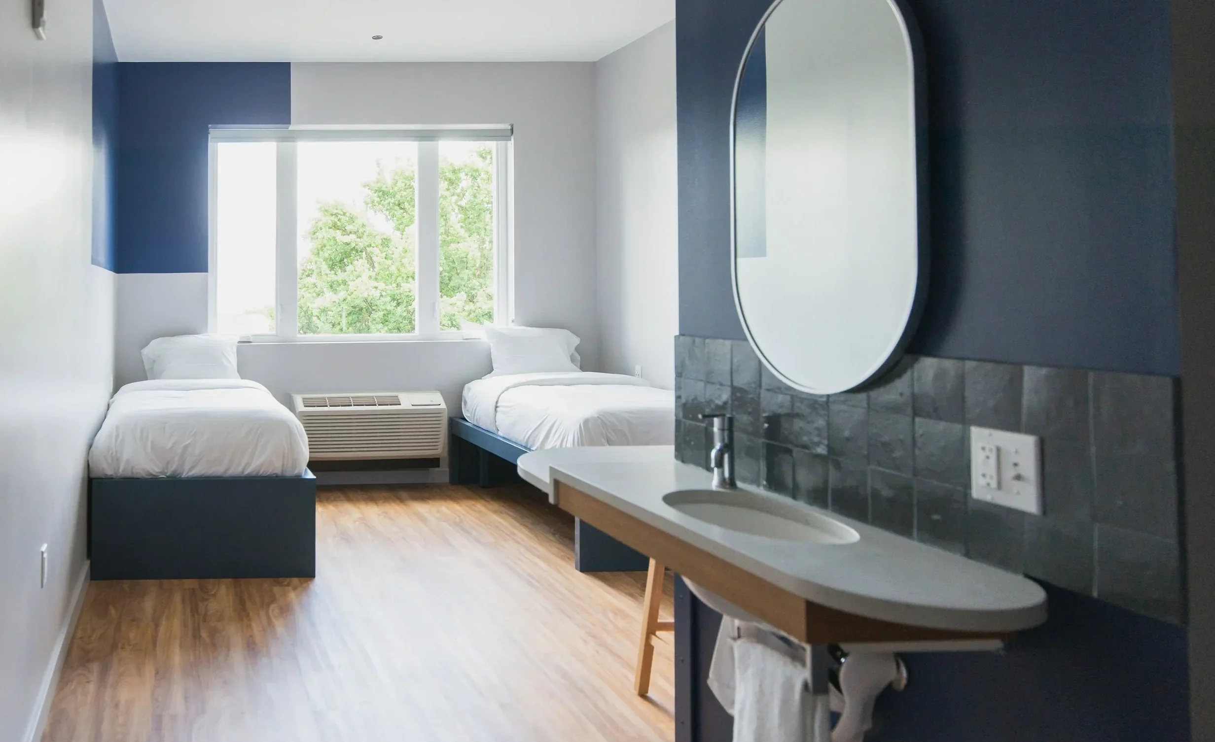 A small, modern hotel room with two twin beds, a window with greenery outside, and a sink with a mirror in the foreground.