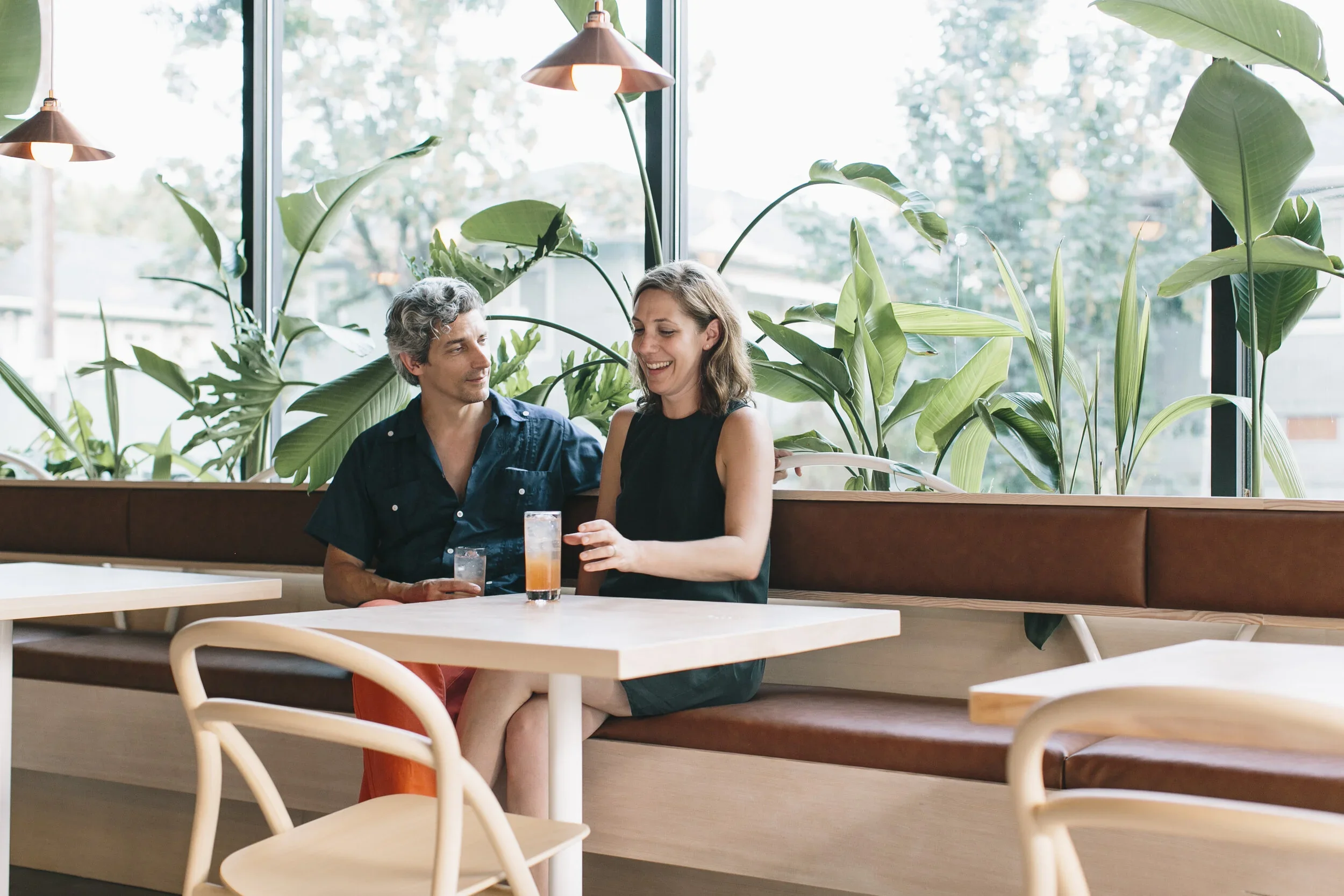 A man and woman sitting at a booth in a cafe with large green plants and a window in the background, sharing drinks and smiling.