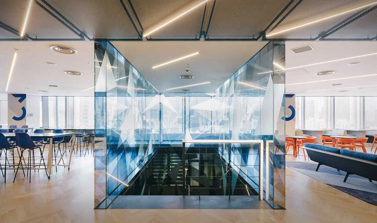 Modern office lobby with glass walls, seating areas, and a staircase leading down, featuring contemporary lighting and city views.