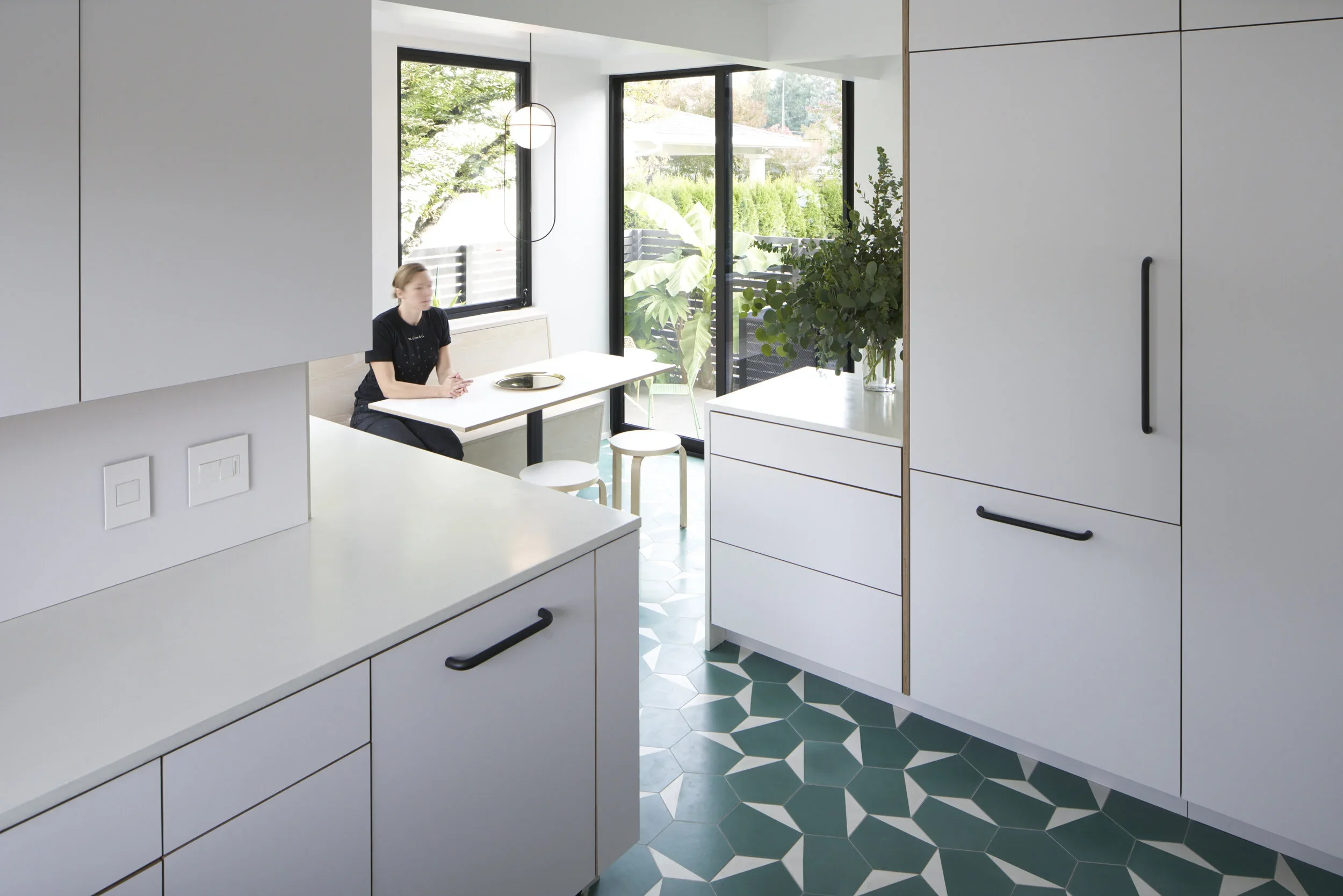 Modern kitchen with white cabinets, black handles, a patterned green and white tile floor, large windows, and a woman sitting at a white dining table near a sliding glass door with a view of greenery outside.