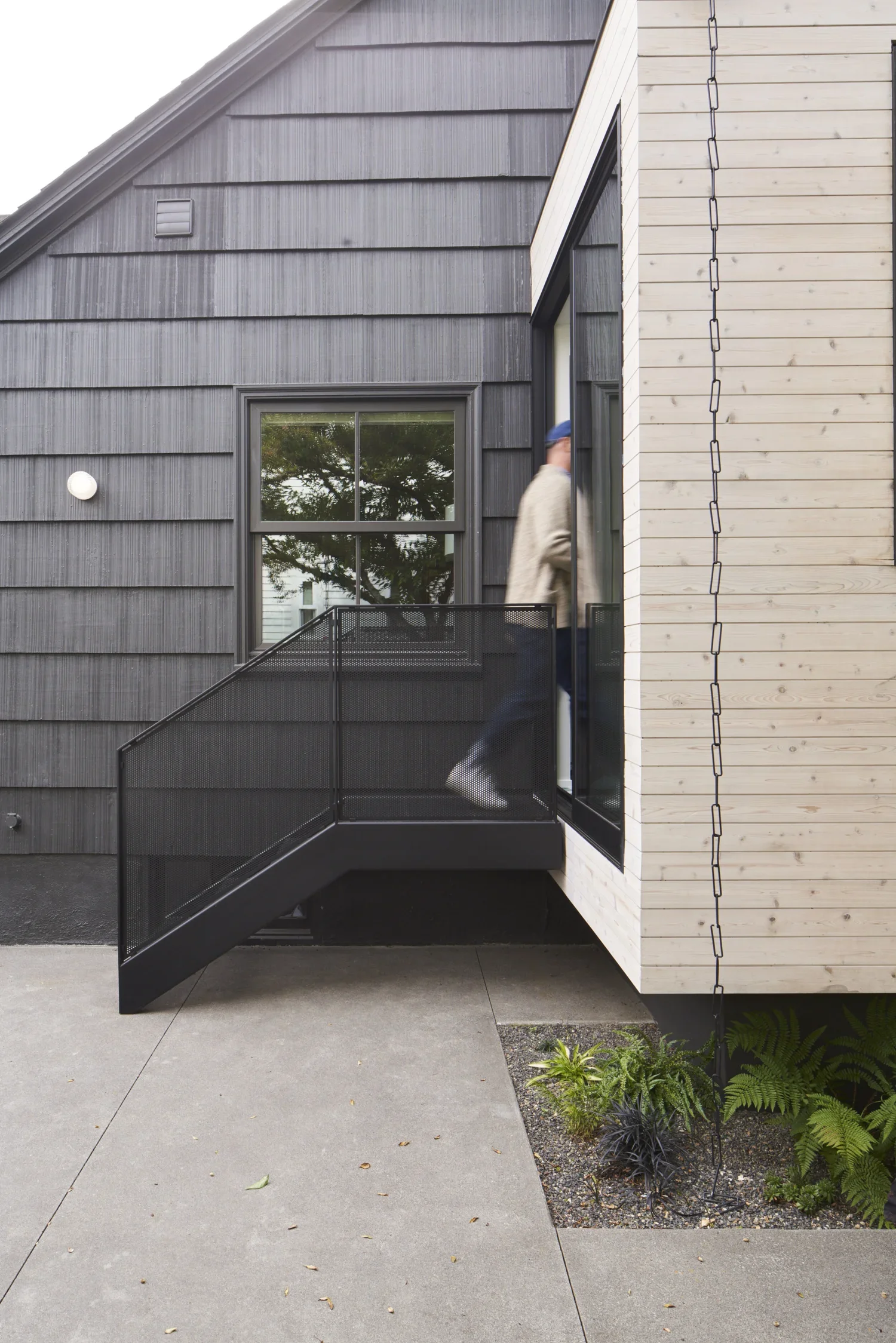 A person walking up small outdoor metal stairs outside a modern house with black and natural wood siding.