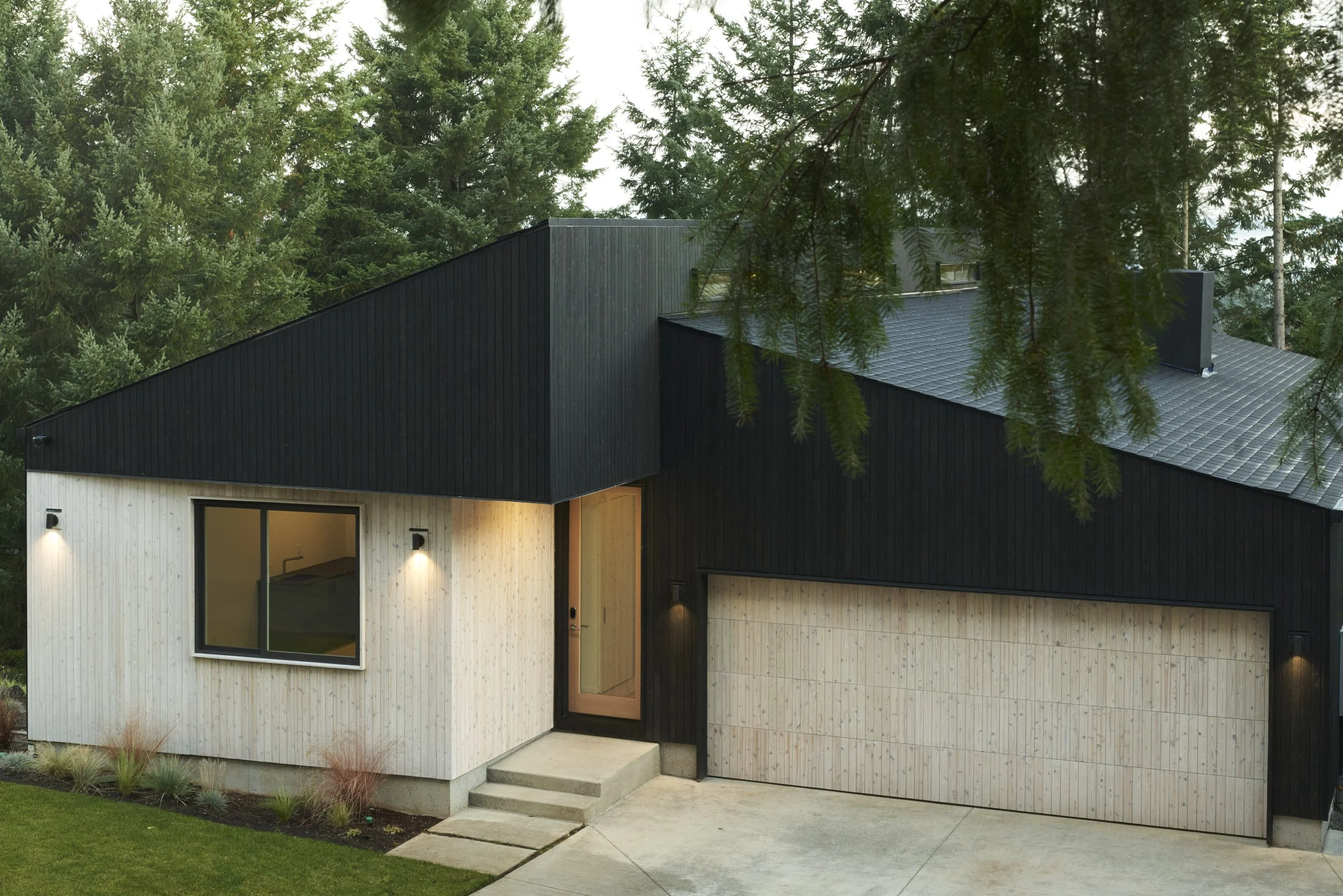 Modern house with black and light wood exterior, large window, attached garage, concrete steps, surrounded by greenery and trees.