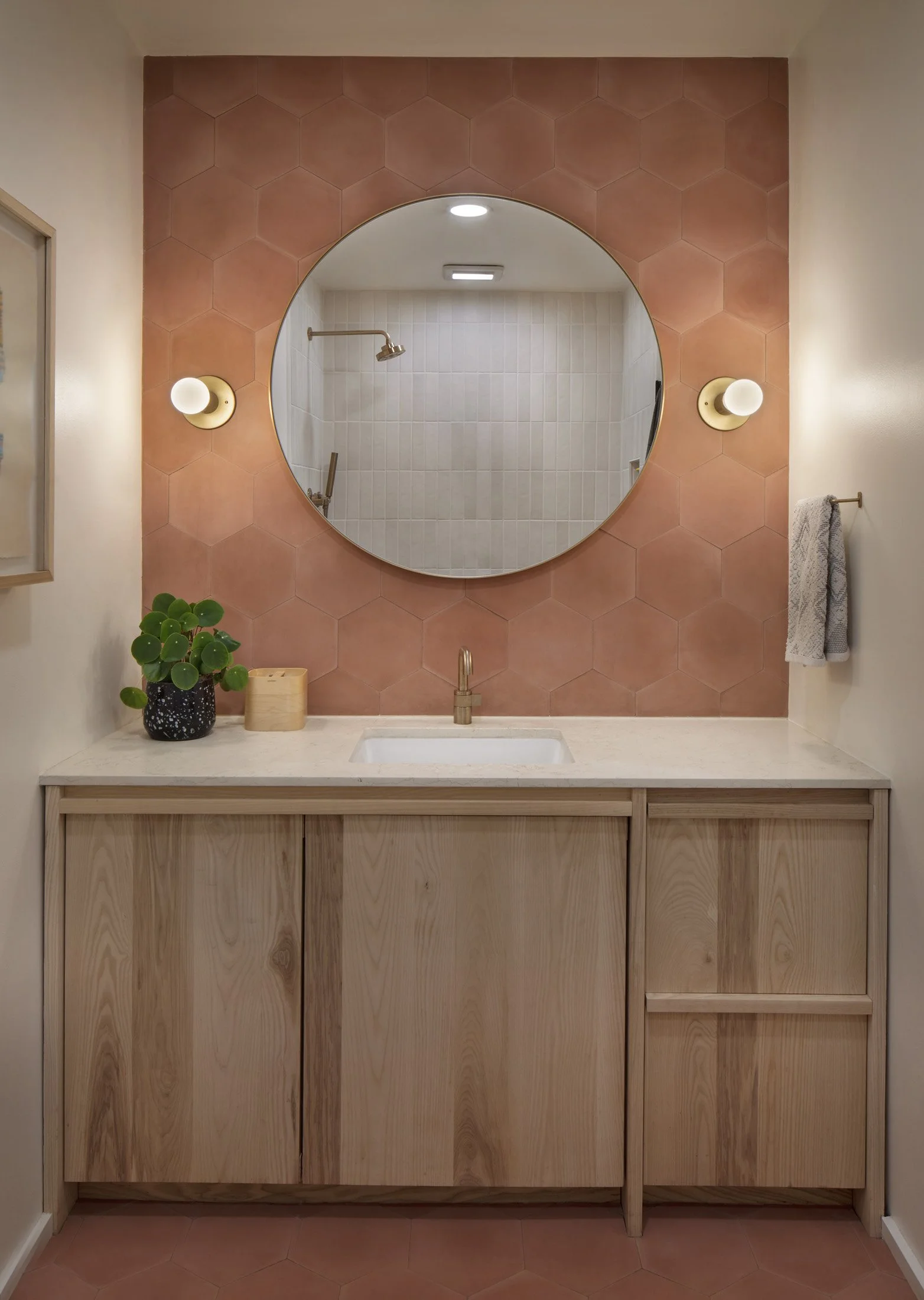 Bathroom vanity with a wooden cabinet, light-colored countertop, round mirror above, pink hexagonal tiled wall, two wall-mounted light fixtures, a small plant, and a towel on a rack.