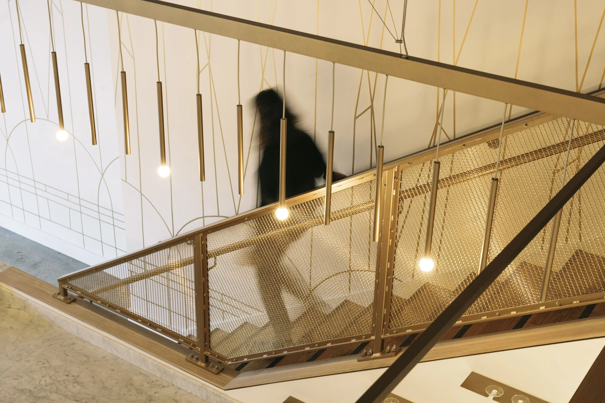 A person with long hair wearing a black top walking up a staircase enclosed by a gold metal railing and gold hanging lights, viewed from above.