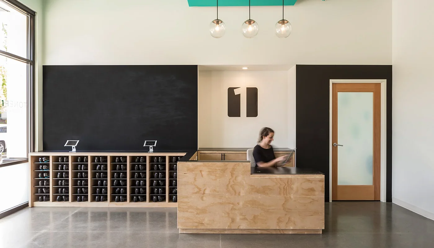 Modern coffee shop counter with a wooden cabinet, a woman working, a black chalkboard wall, and contemporary lighting fixtures.