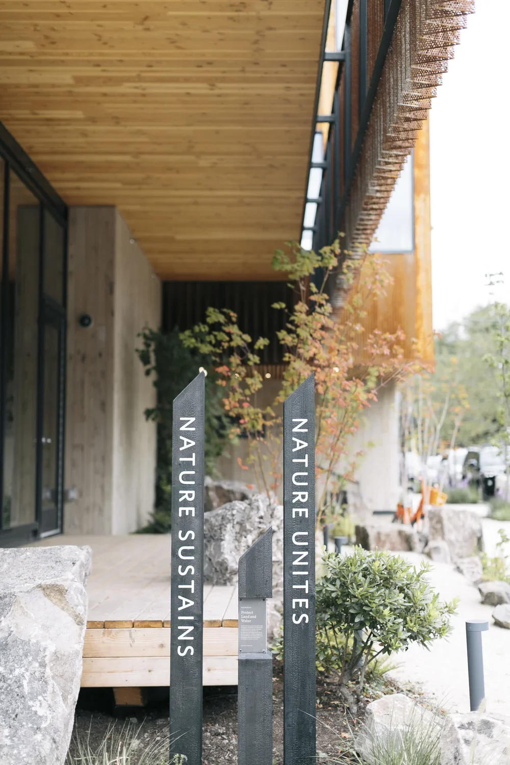Outdoor modern building with wooden roof and black signs labeled "Nature Sustains" and "Nature Untes," with bushes, rocks, and trees nearby.