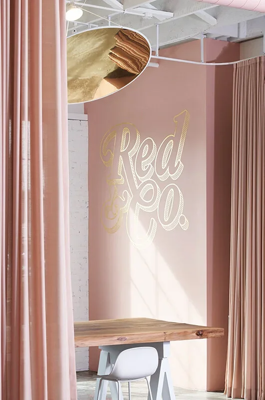 Interior of a space with pink walls, pink curtains, a wooden table, a white chair, and a wall with the logo 'Red co.' in large gold script, with a mirror or shiny fixture overhead reflecting part of the room.