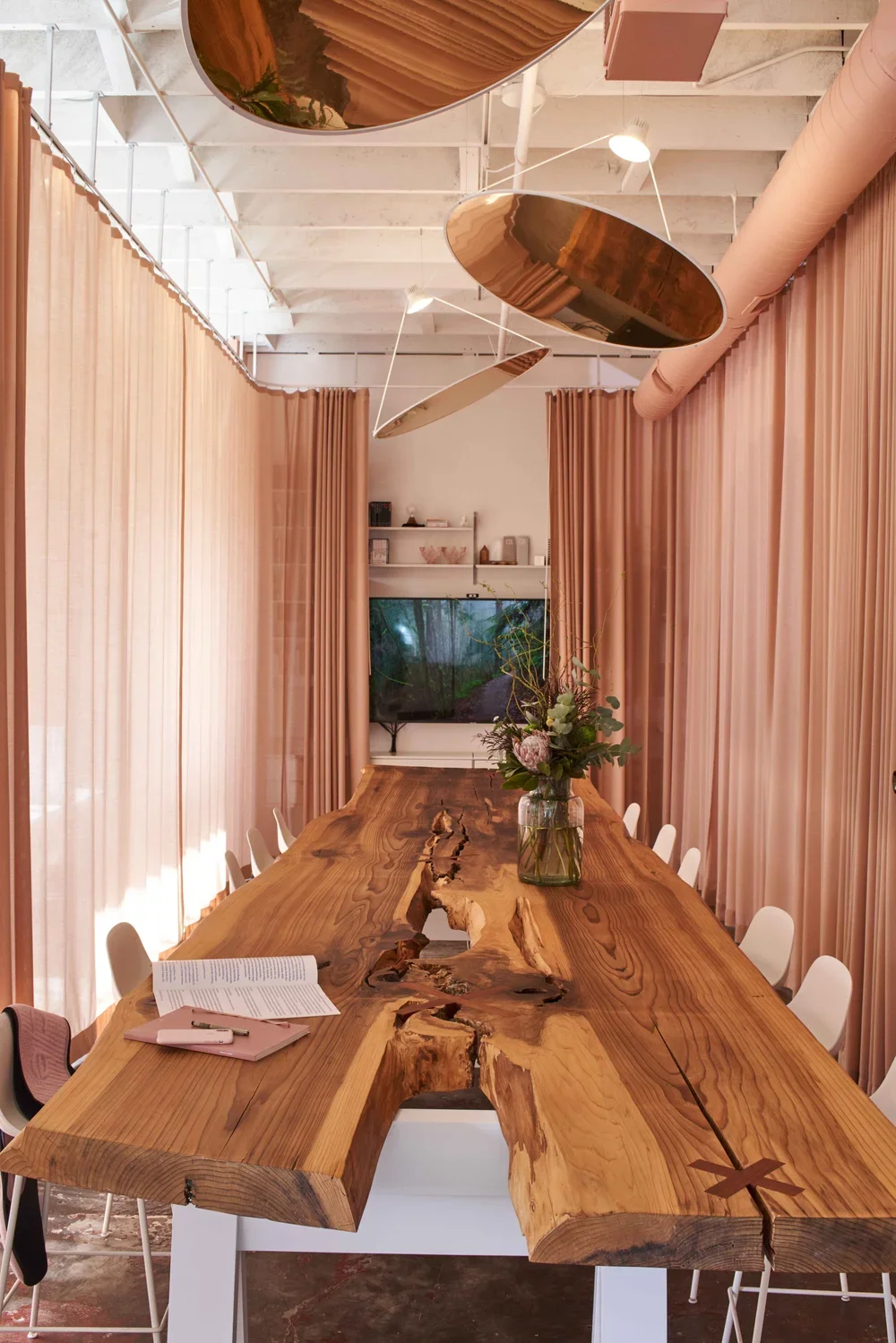 Modern dining room with a large, natural wood table with a live edge and knots, surrounded by white chairs, pink curtains, botanical flower arrangement in a glass vase, and a flat-screen TV on the wall in the background.