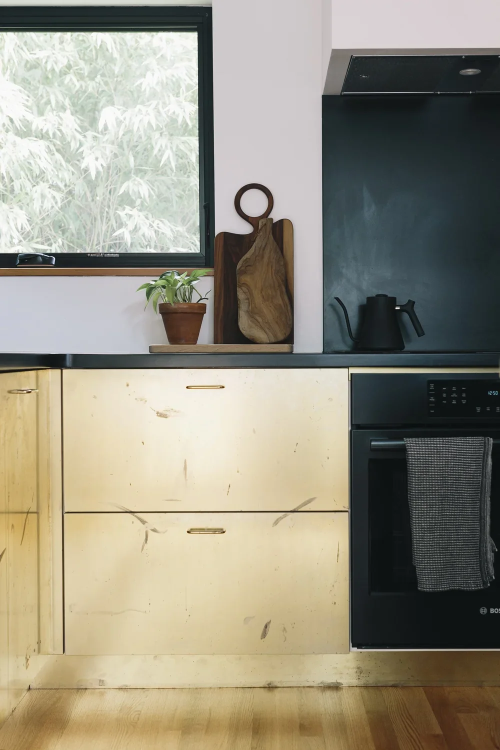 Kitchen countertop with potted plant, wooden and cutting boards near a window, black kettle, and oven with a dish towel hanging from the handle.