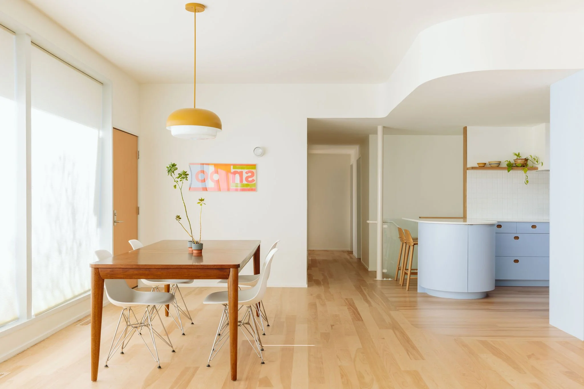 Minimalist dining area with wooden table, four white chairs, potted plants, and a yellow pendant lamp, adjacent to a kitchen with blue cabinetry and wooden shelves, in a bright, airy home.