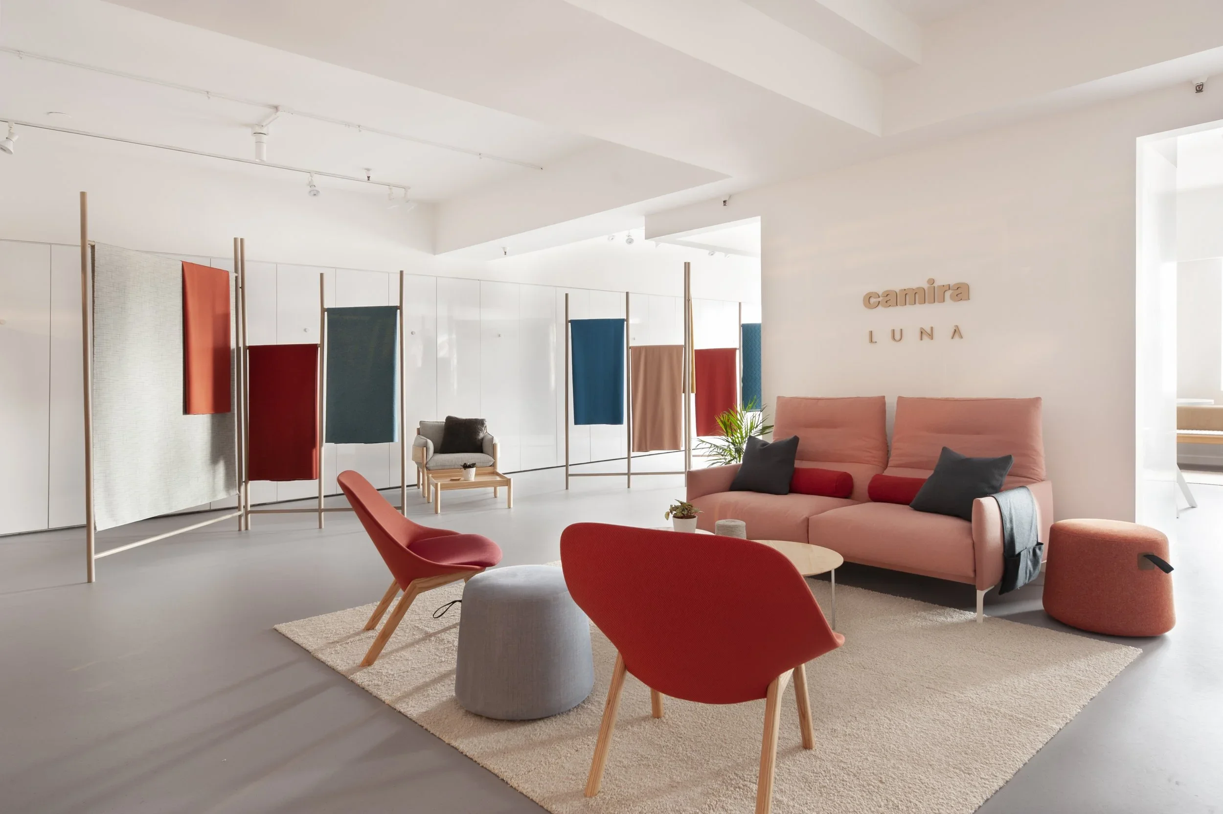 Modern interior with pink sofa, red chairs, colorful hanging fabric panels, and the signs 'Camira' and 'LUNA' on the wall.