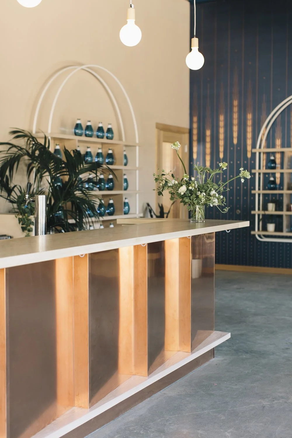 Modern cafe interior with wooden and metallic bar counter, hanging lights, a floral arrangement on the counter, and shelves with blue jars in the background.