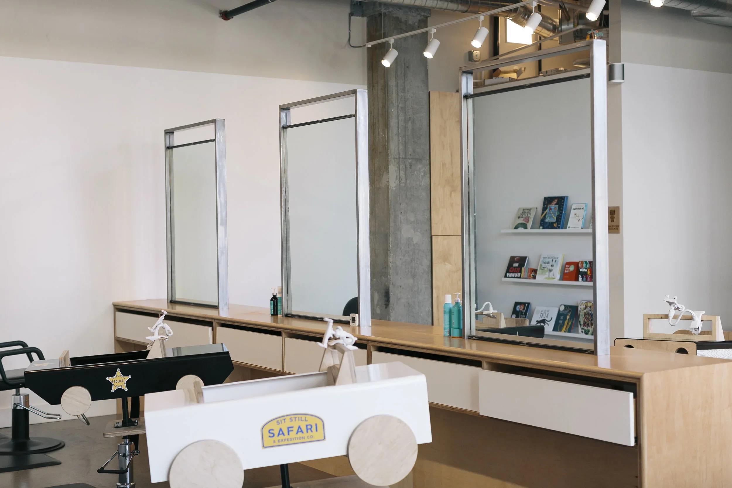 Interior view of a modern salon or grooming station with three wooden work stations separated by metal dividers, a bookshelf with children's books, and a cart with supplies including hand sanitizer and disinfectant spray.