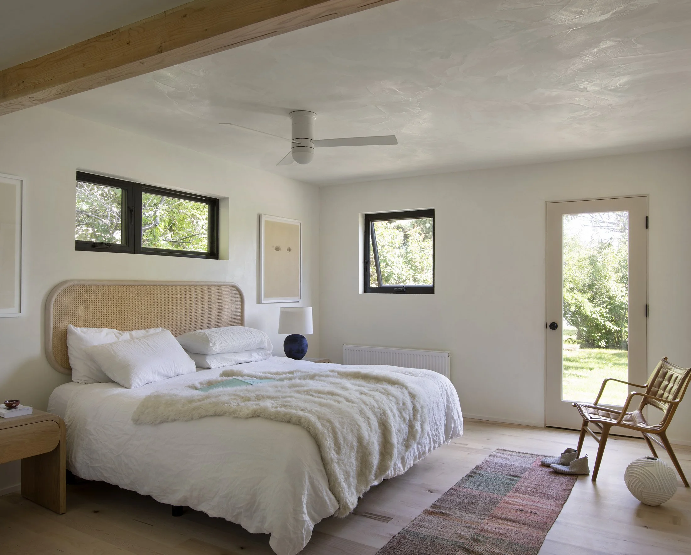 Bedroom with white walls, a large bed with white bedding, a wicker headboard, a wooden side table with a small item, a blue and white table lamp, two small windows, a glass door leading outside, a wicker chair with slippers, a striped rug on light wood floor, and a ceiling fan.