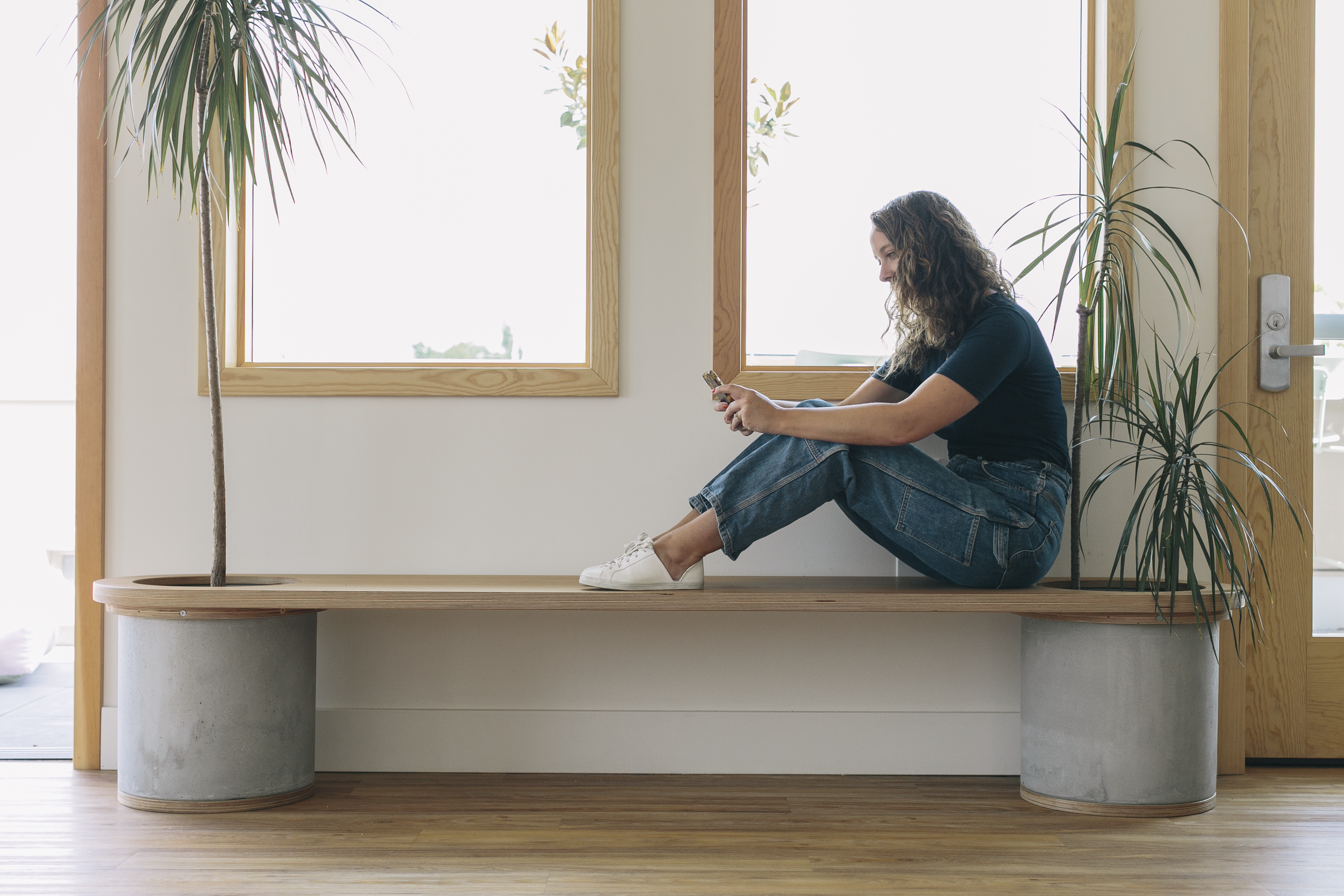 A woman with curly hair wearing a dark shirt, jeans, and white sneakers sitting on a wooden bench with concrete supports in a bright room with large windows, holding and looking at her phone.