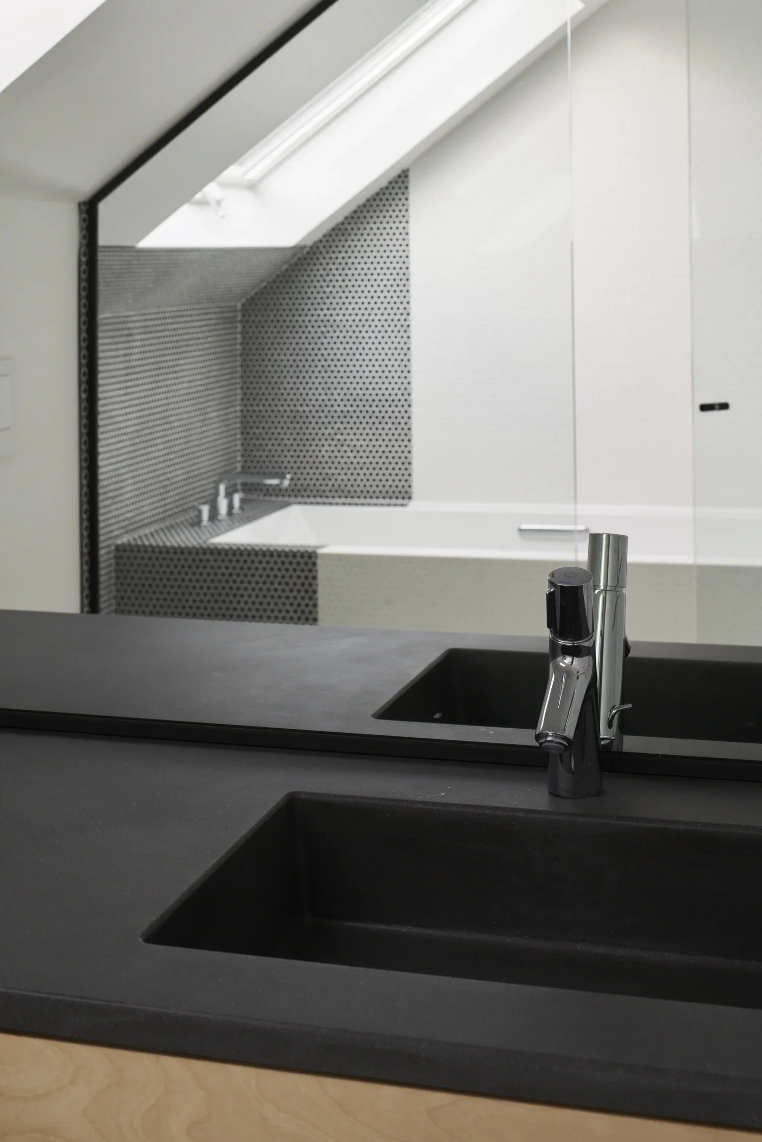 Modern kitchen with black countertop, double black sinks, and chrome faucet, overlooking a bathroom with a bathtub and patterned black and white tiled walls.