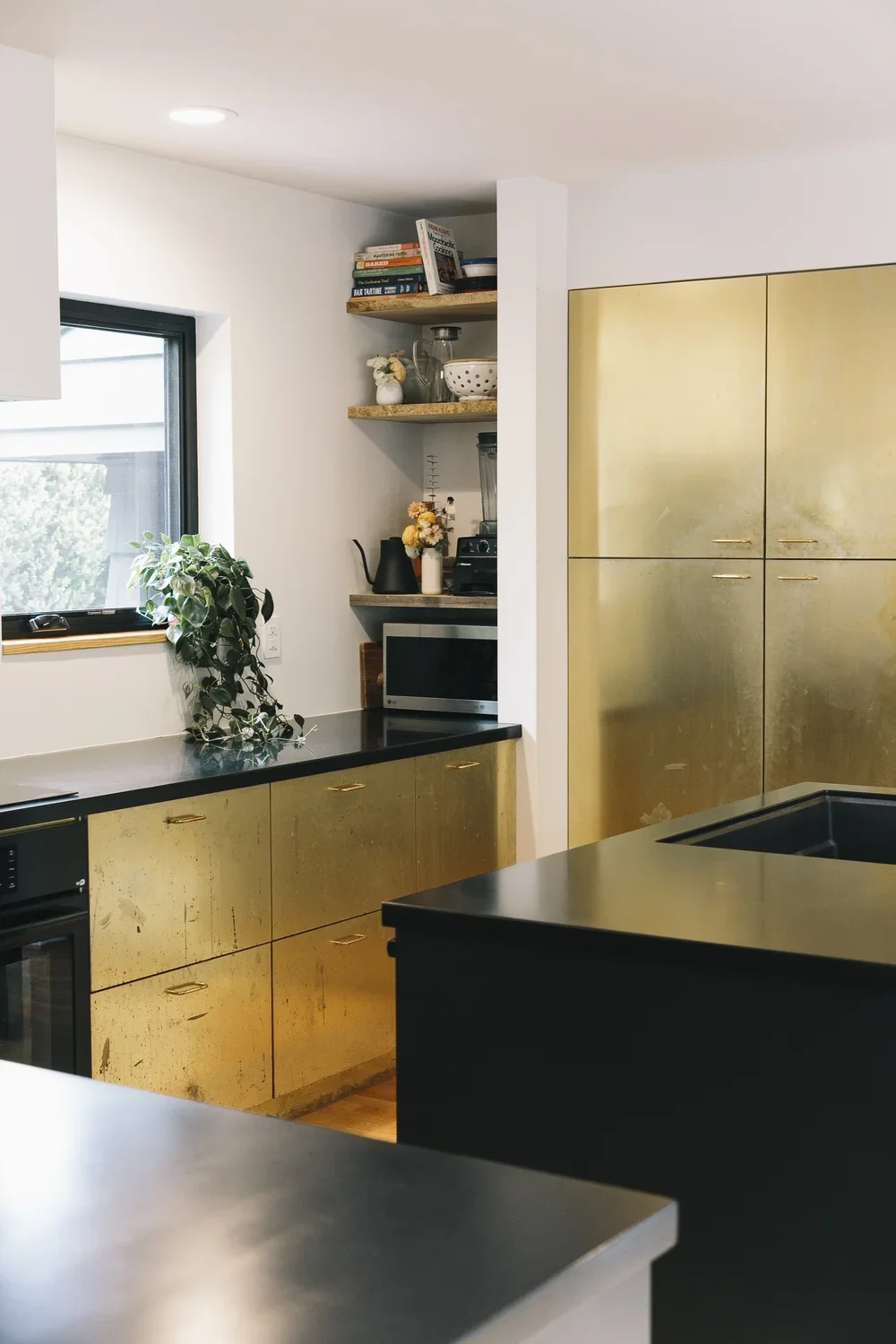 Kitchen with black and gold cabinets, a window with a plant on the counter, open shelves with books and kitchen items, and a microwave.