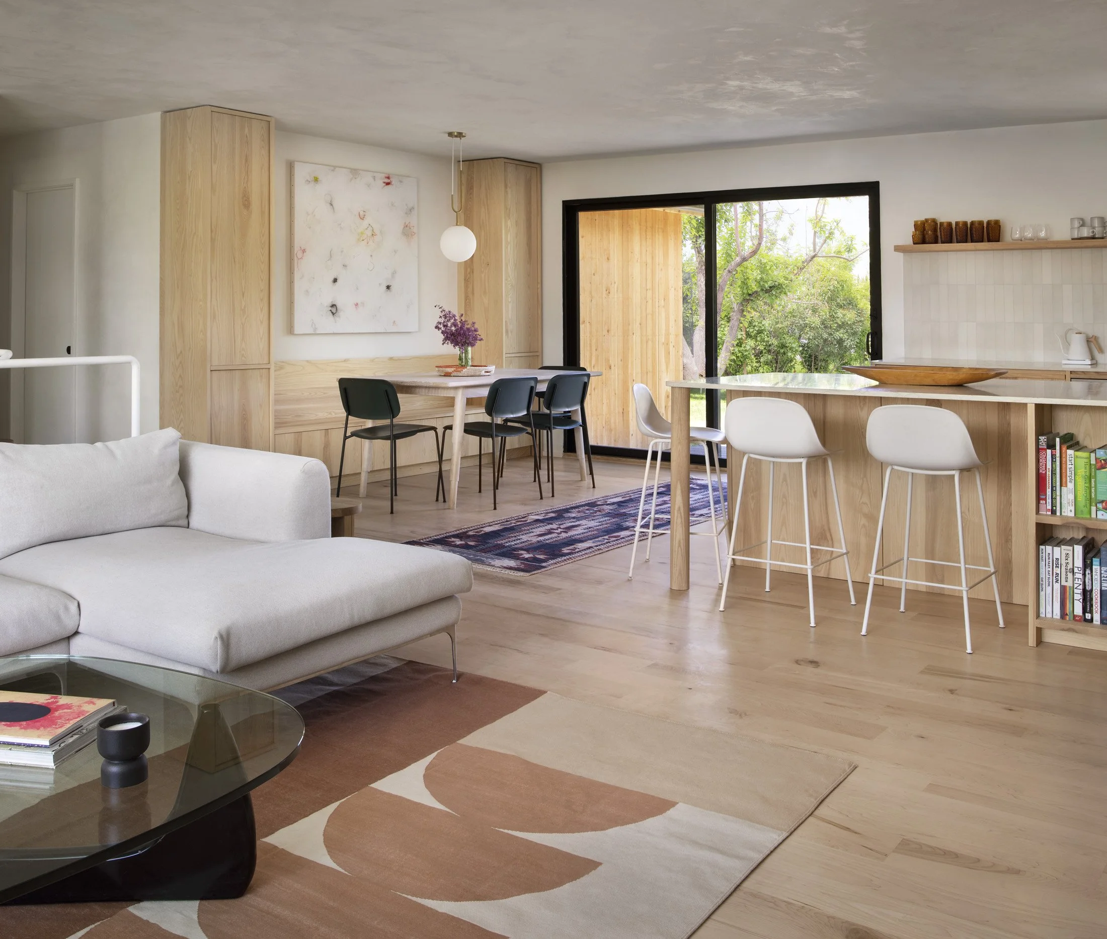Open-concept living and dining area with wooden accents, white sofa, black and white chairs, a large window, and a bookshelf.