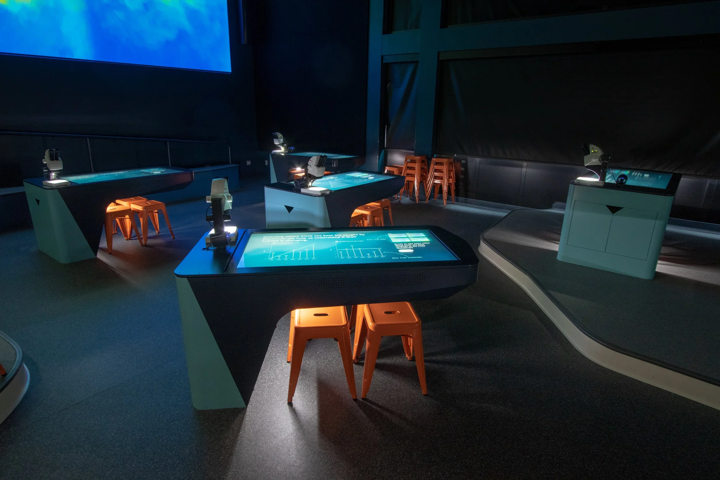 Interactive science exhibit with multiple digital touch screens and microscopes in a dark room with blue lighting.