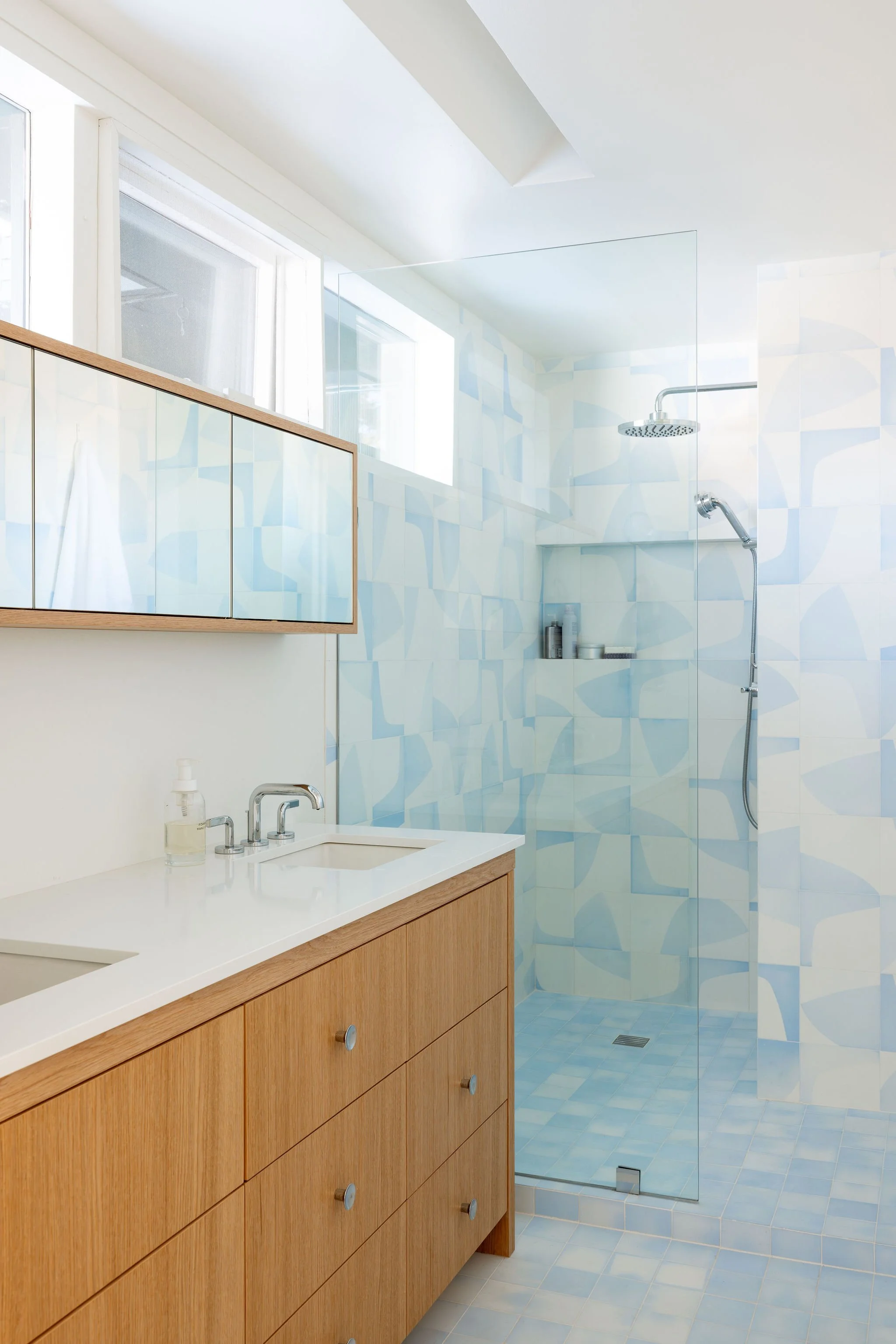 Modern bathroom with wooden vanity, white countertop, and a glass-enclosed shower with blue patterned tiles and built-in shelf.