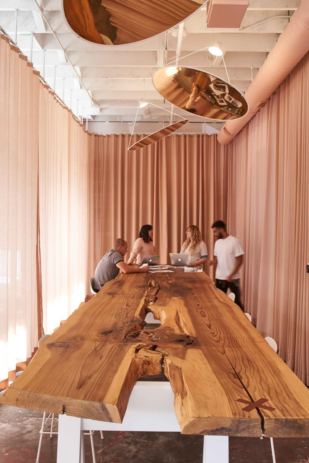 A large wooden conference table with a natural, rustic finish, surrounded by people working on laptops in a room with pink curtains and modern ceiling lights