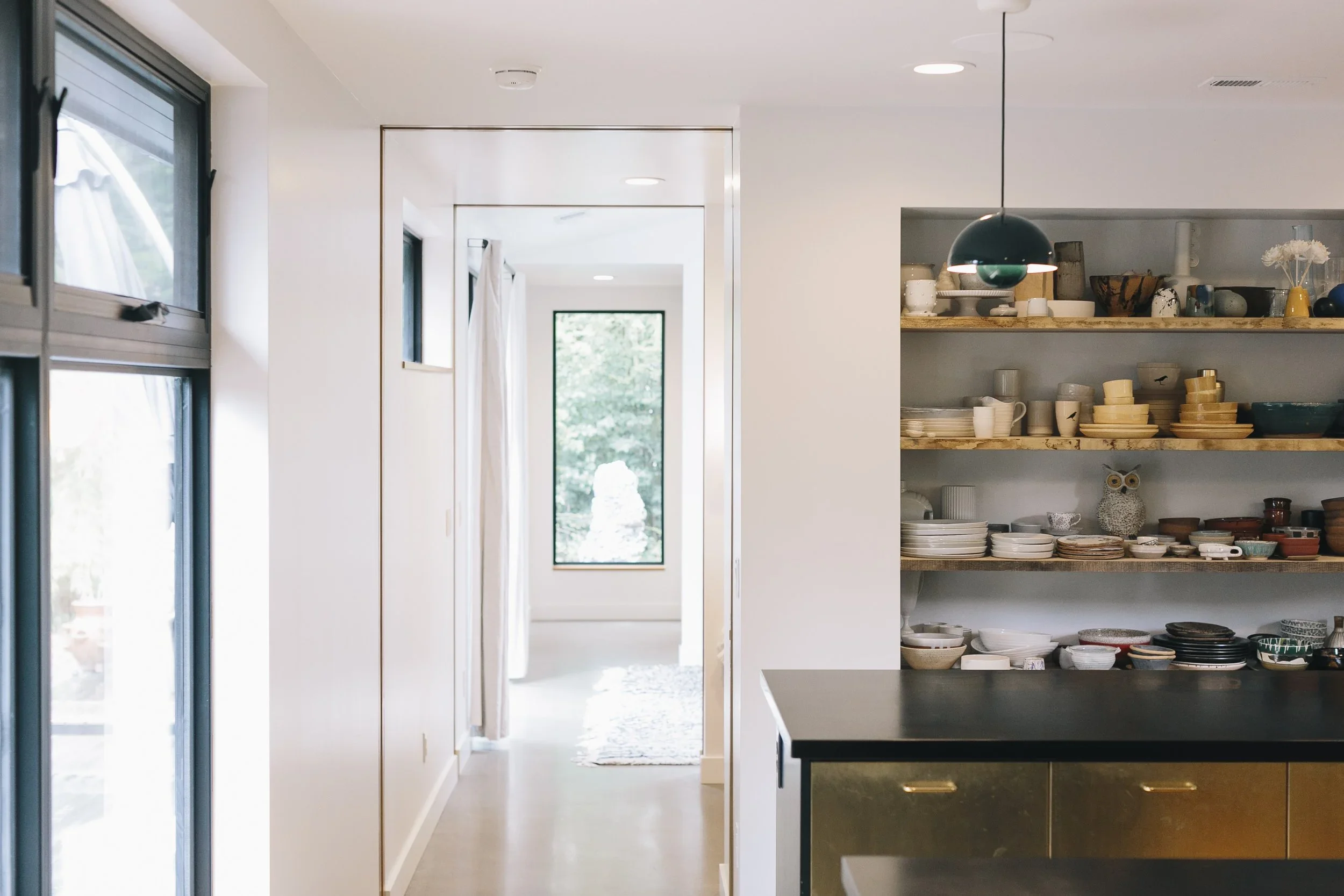 A modern kitchen with open shelving displaying plates and bowls, a black countertop, and a pendant light. Sunlight comes through windows, illuminating a hallway leading to a bright room.
