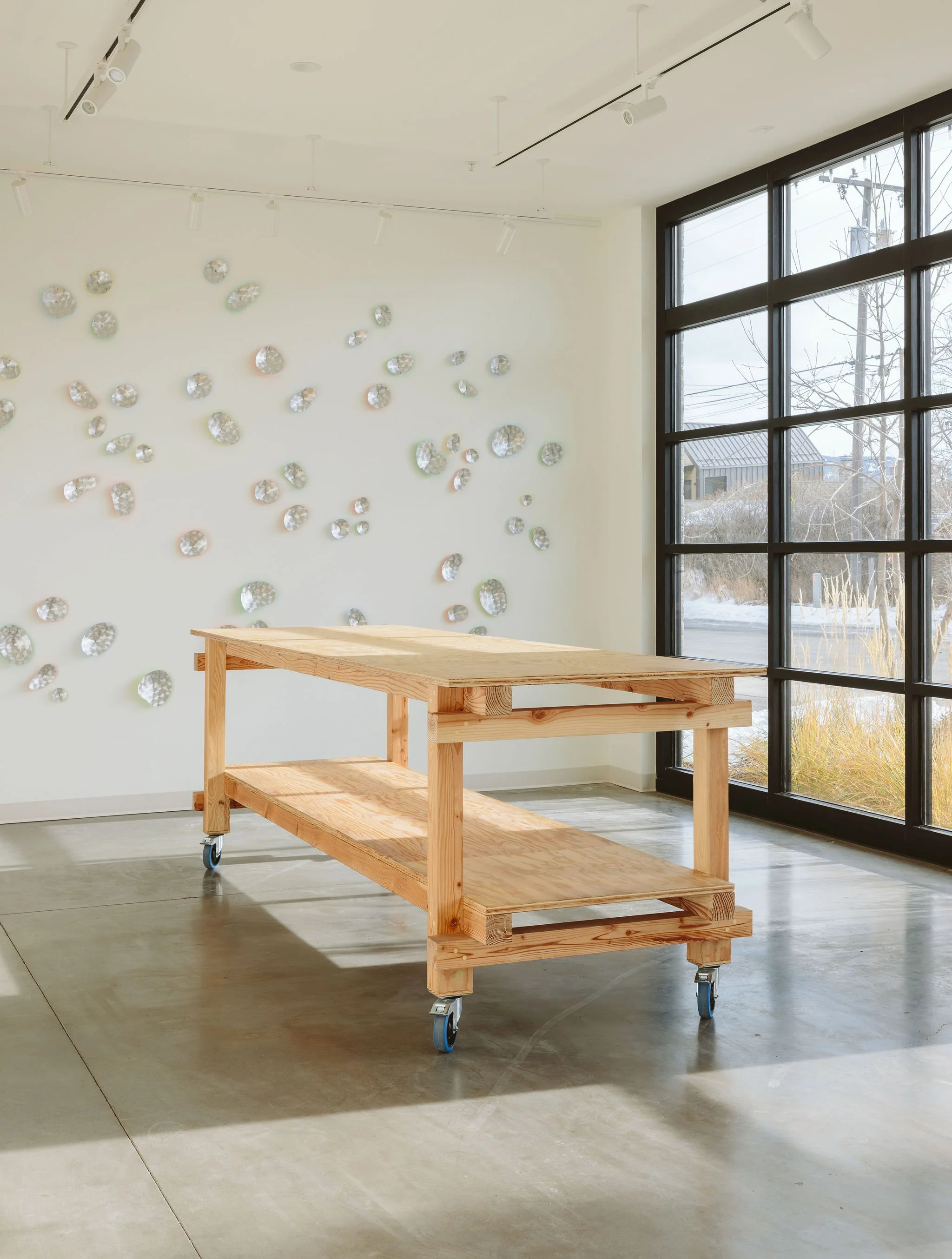 Empty wooden cart on wheels inside a modern art gallery with white walls, decorative silver circular wall art, and large windows showing a snowy outdoor scene.