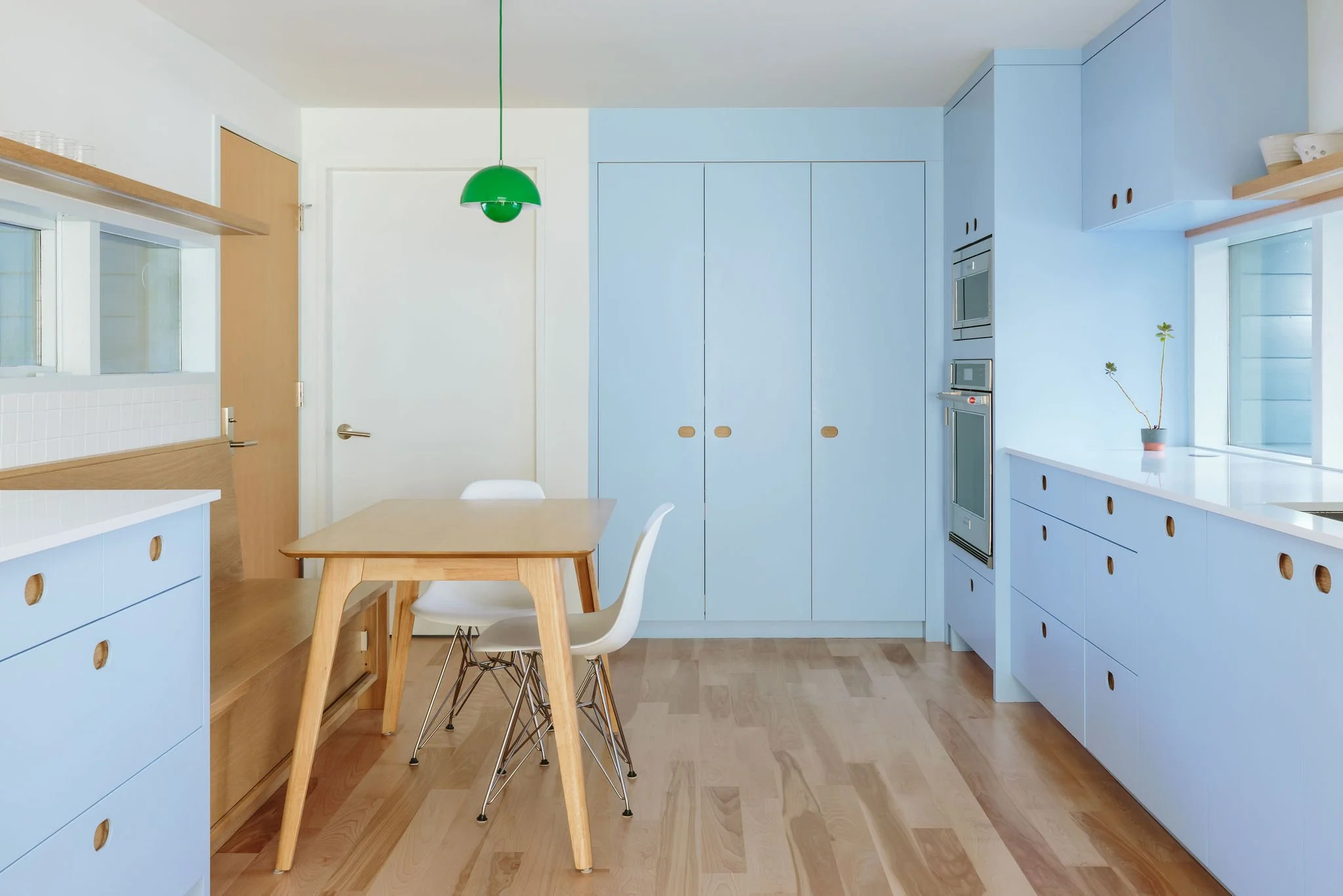 Modern kitchen with light blue cabinetry, wooden flooring, a wooden dining table with two white chairs, and a green pendant light hanging above the table.