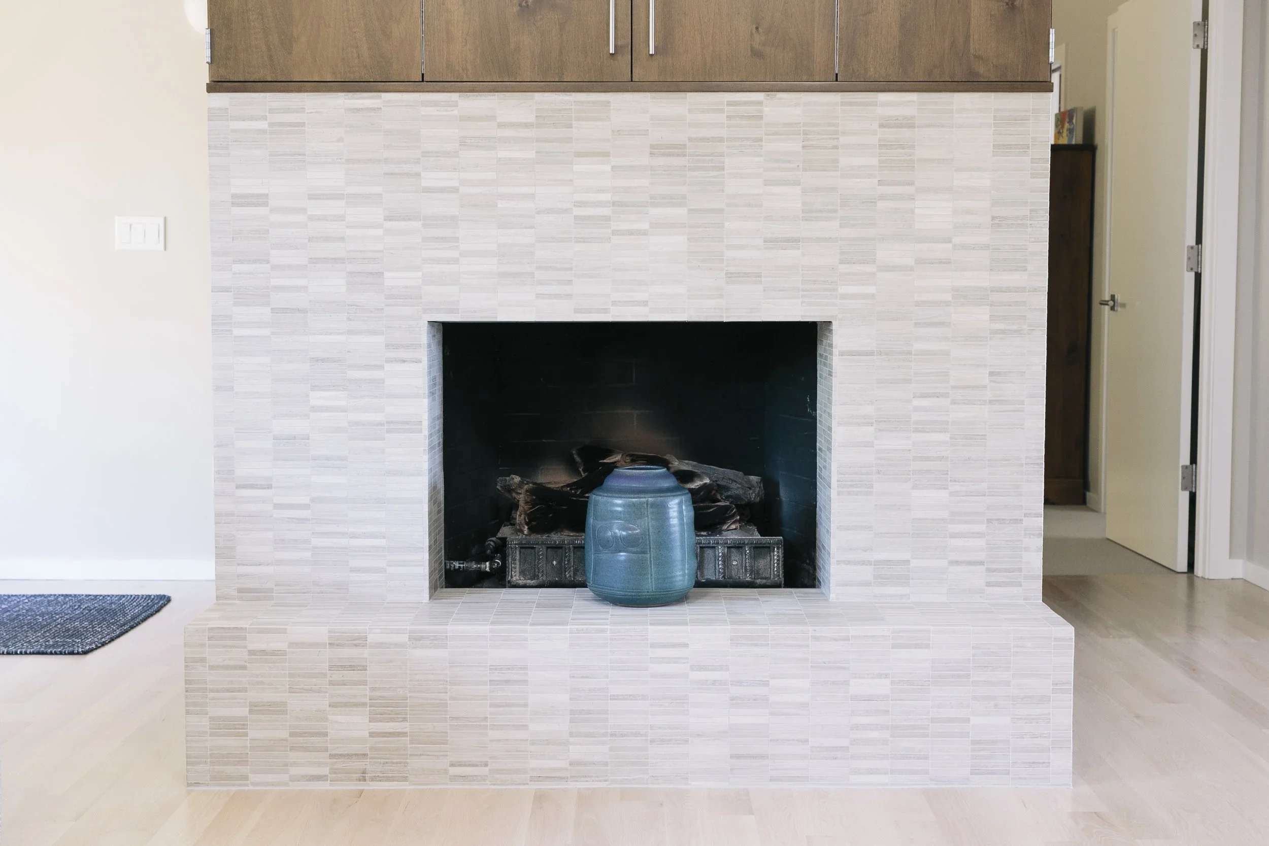 Modern fireplace with beige tile surround and a blue ceramic pot on the hearth, in a bright room with light-colored wooden floors and a partial view of an open door.