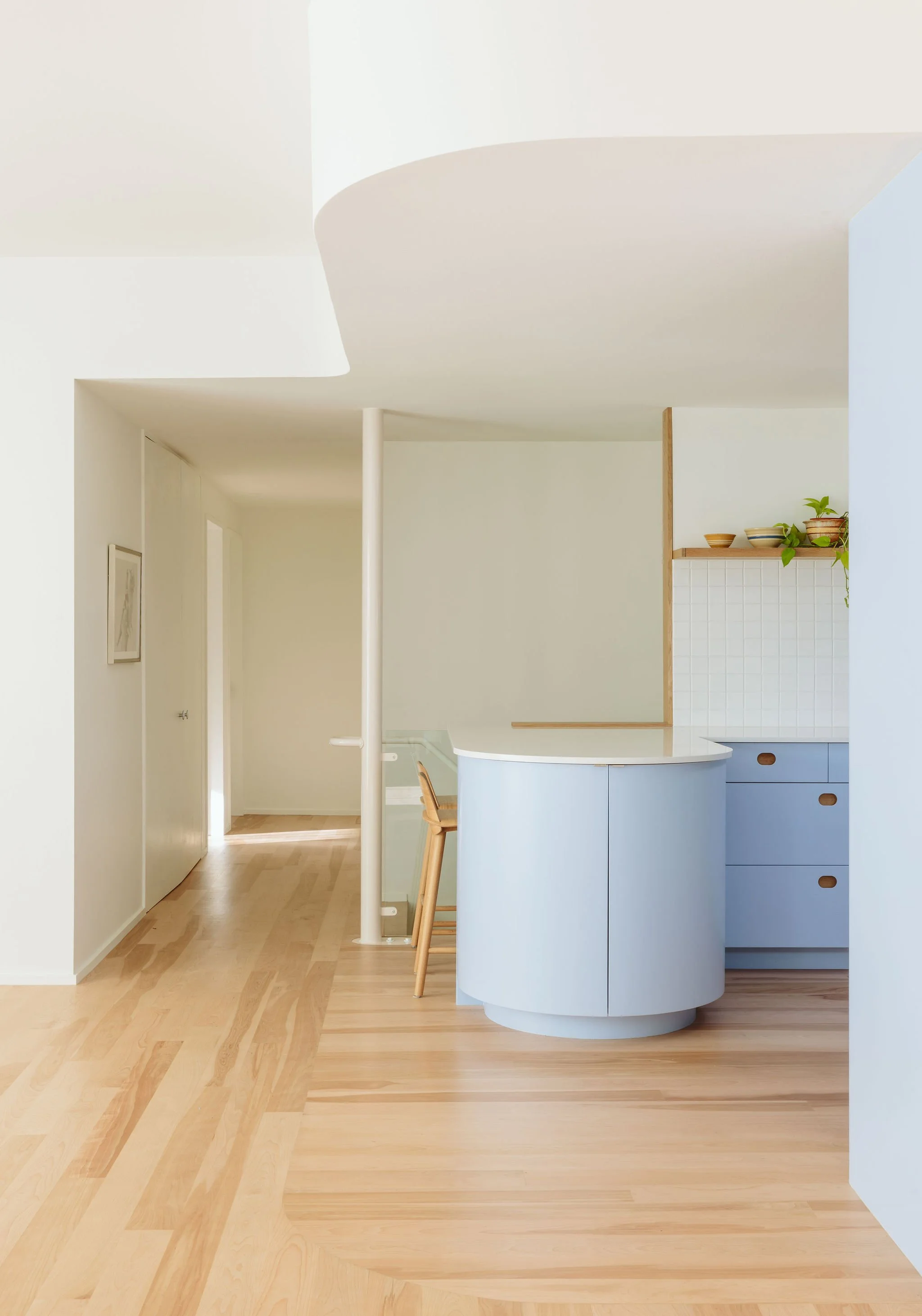 Minimalist kitchen with pale blue cabinetry, white countertops, and open shelving with small potted plants. Light wood floors and white walls with soft natural light.