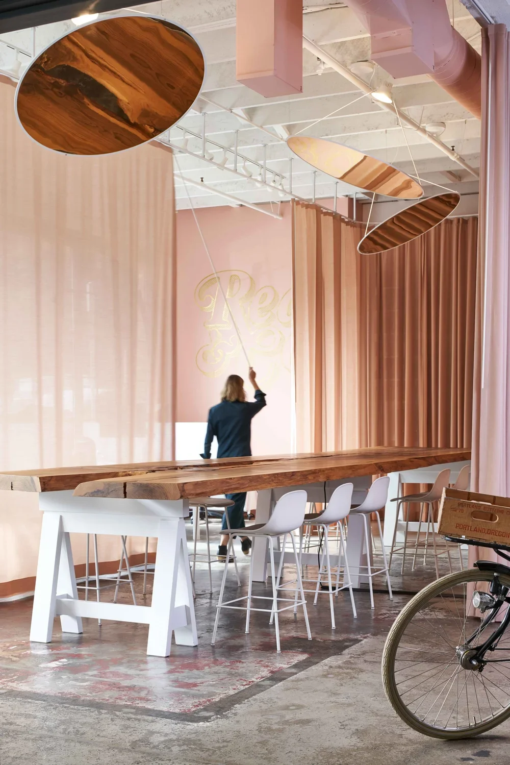 Interior of a modern space with pink curtains, a high wooden table with white chairs, abstract hanging art pieces, and a person with long hair throwing a rope. A bicycle with a wooden crate attached is partially visible in the foreground.