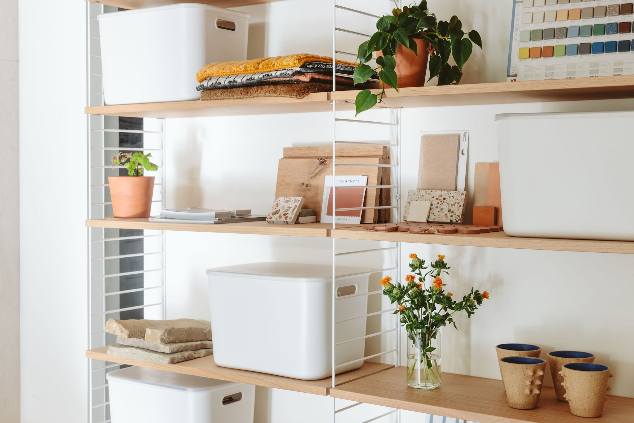 Wooden shelves with potted plants, fabric samples, folders, storage boxes, clay tiles, and decorative cups in a room.