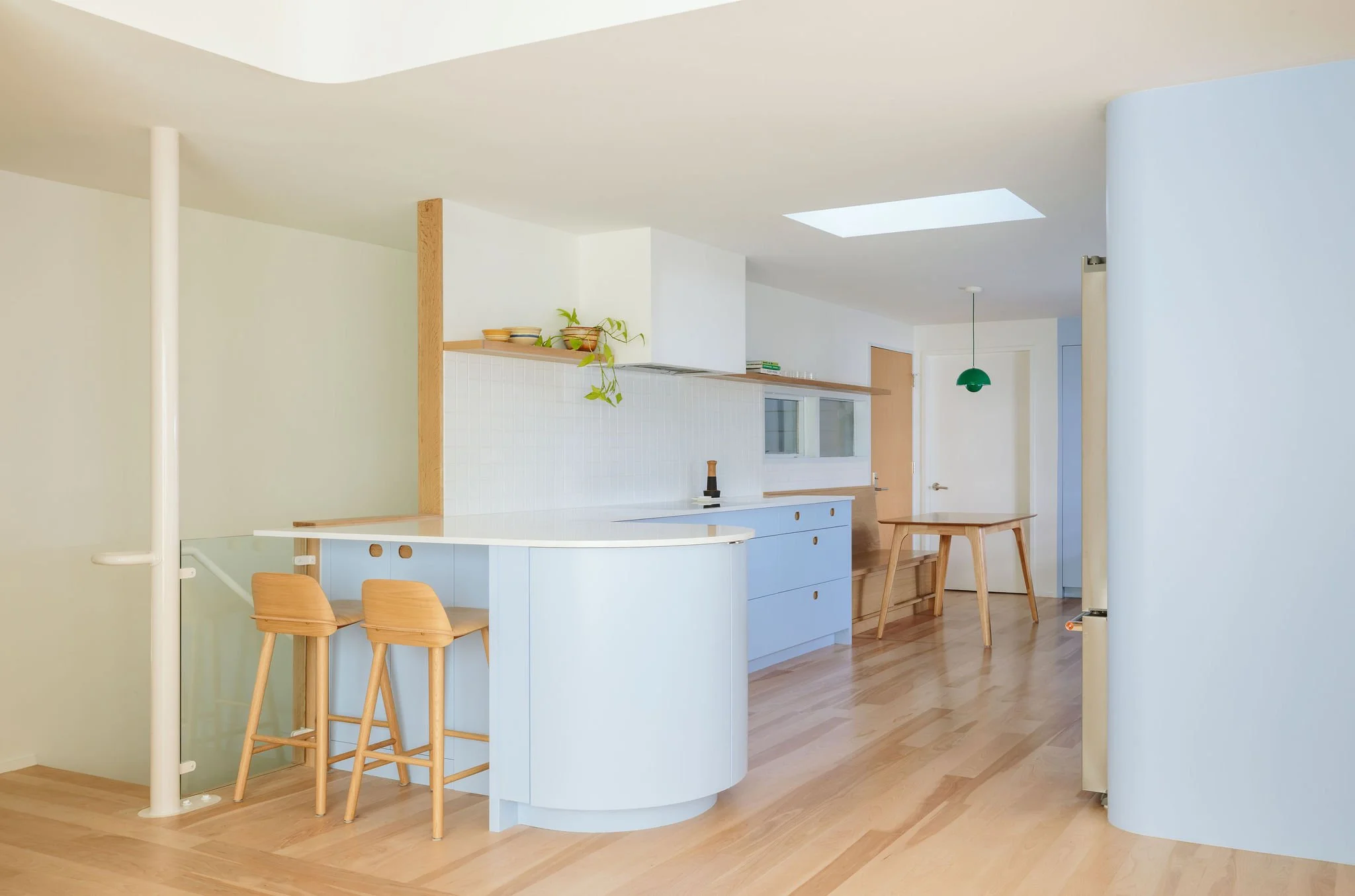 Modern kitchen with light wood flooring, white curved kitchen island, blue cabinets, wooden bar chairs, and a dining table, with a skylight and a green pendant light.