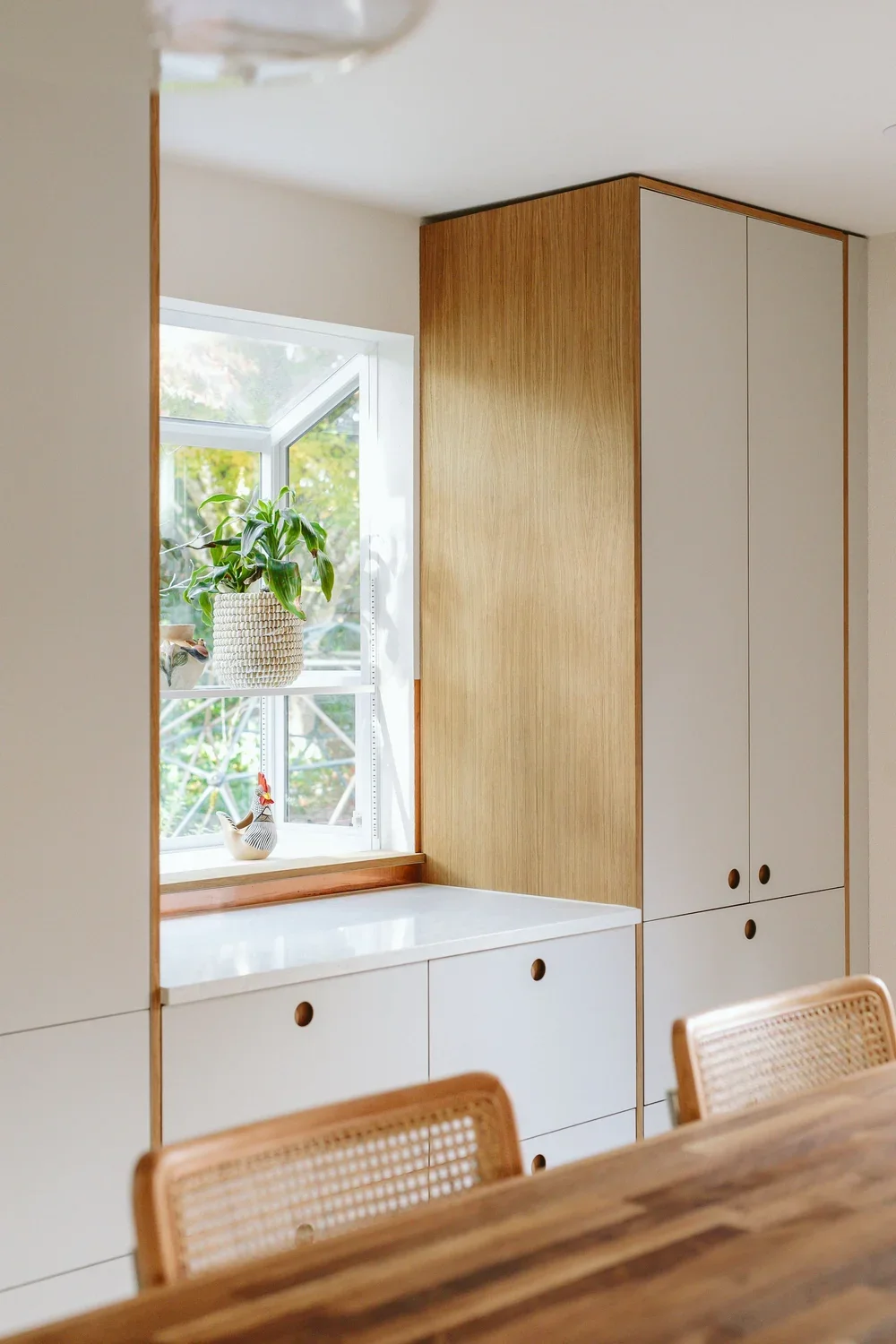 Interior view of a room with a built-in wooden and white cabinet, a window with a potted plant and decorative chicken figurine, and a wooden dining table with rattan-backed chairs.