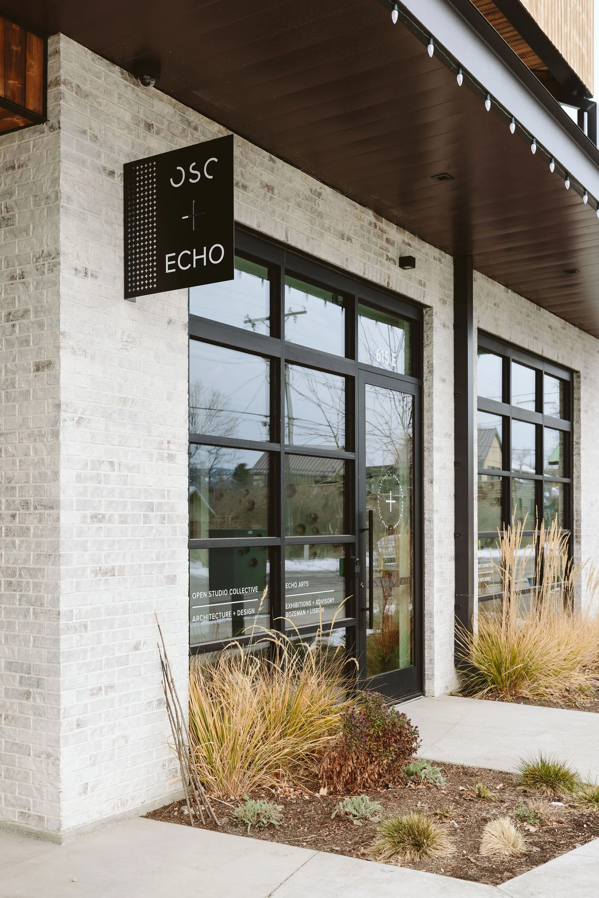 Exterior view of a modern commercial building with a black sign that reads 'JSC + ECHO'. Large glass windows and doors with white lettering display information about the business, including open studio collective, echo arts, architecture and design,