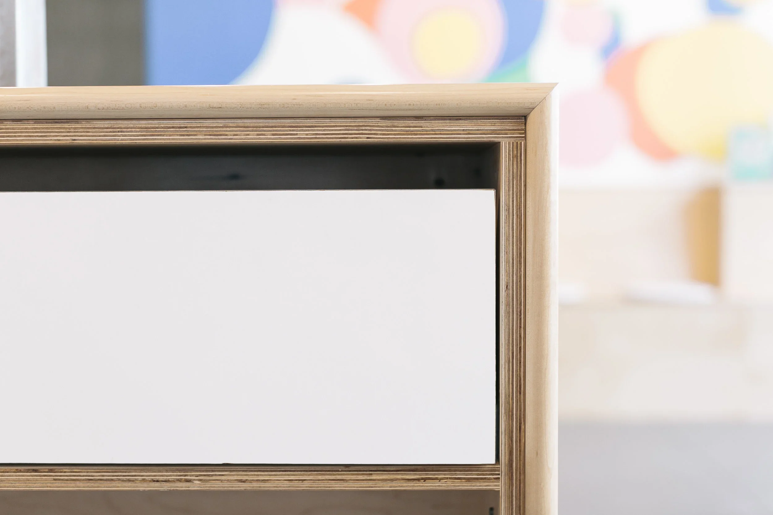 Close-up of a wooden desk with a white drawer, with a colorful abstract painting in the background.
