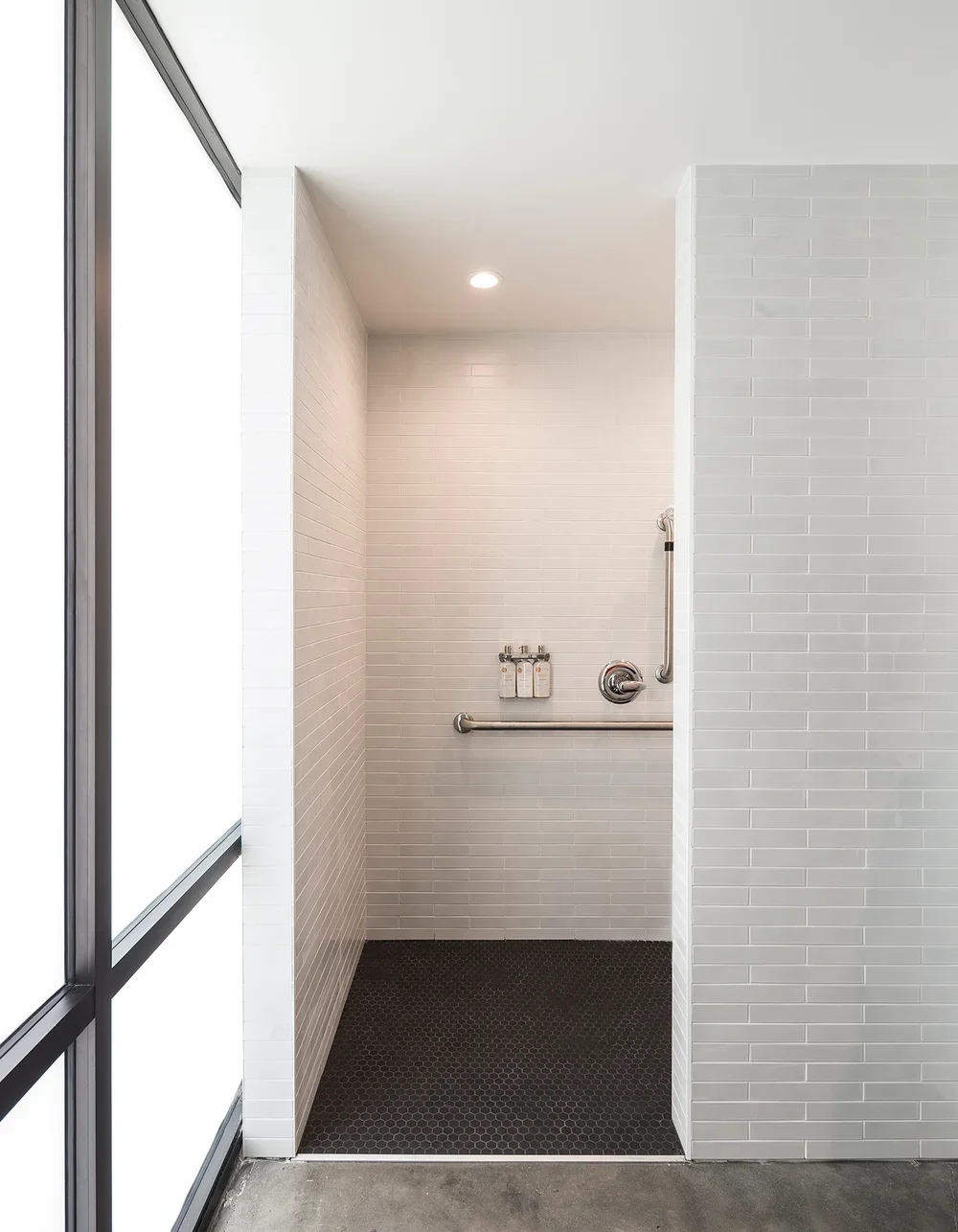Empty accessible shower with white tile walls, black hexagon floor tiles, and a grab bar, located next to a large window with frosted glass.