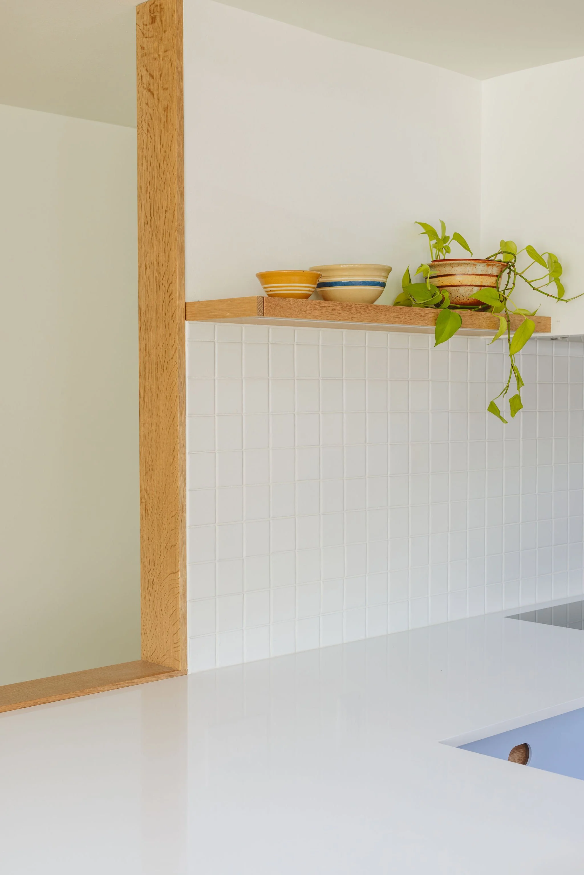 Corner of a kitchen with a white tiled wall, a white countertop, and a wooden shelf with bowls and a potted plant.