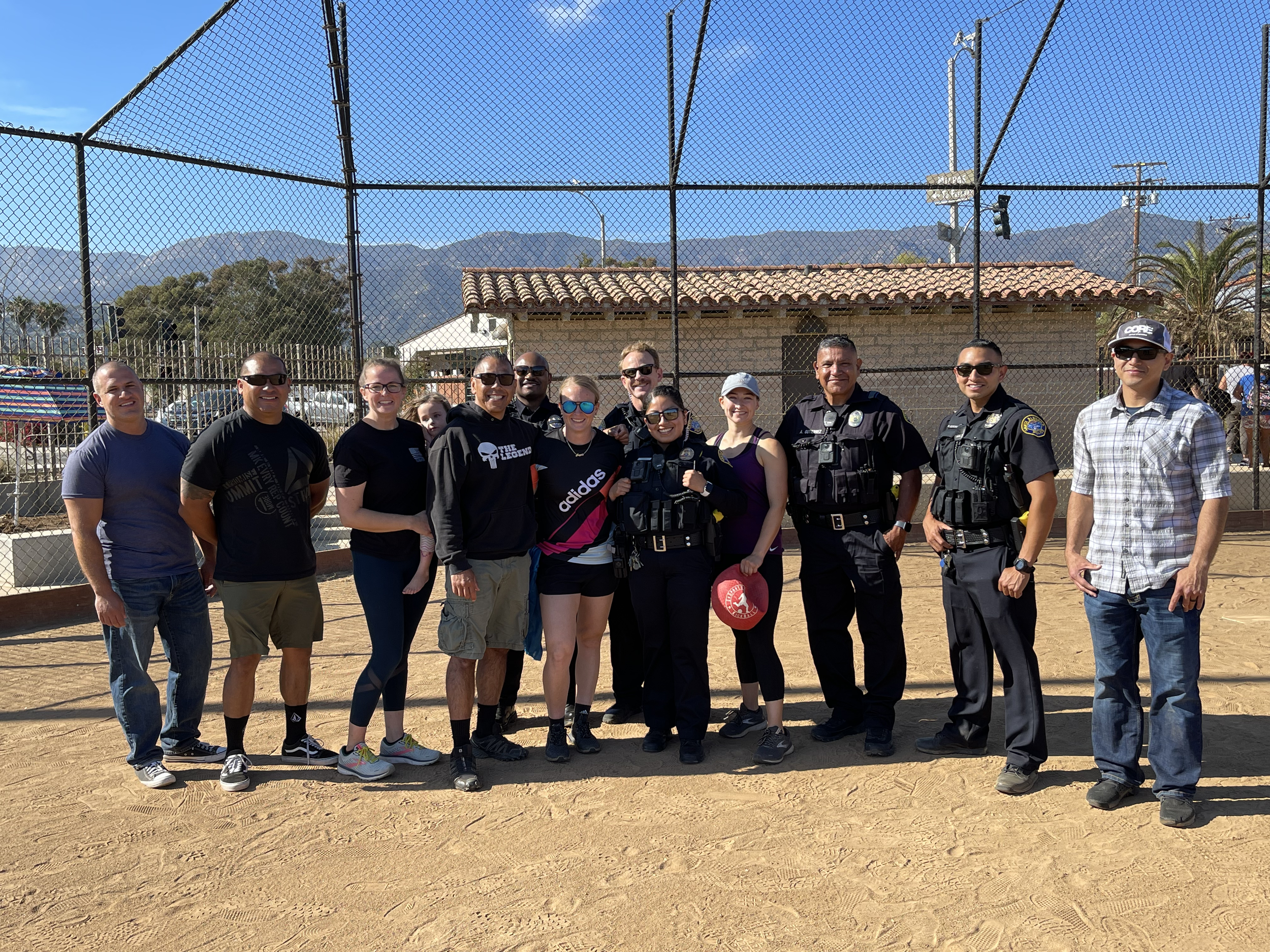 A group of people, including police officers, standing together on a baseball field with mountains and a clear blue sky in the background.
