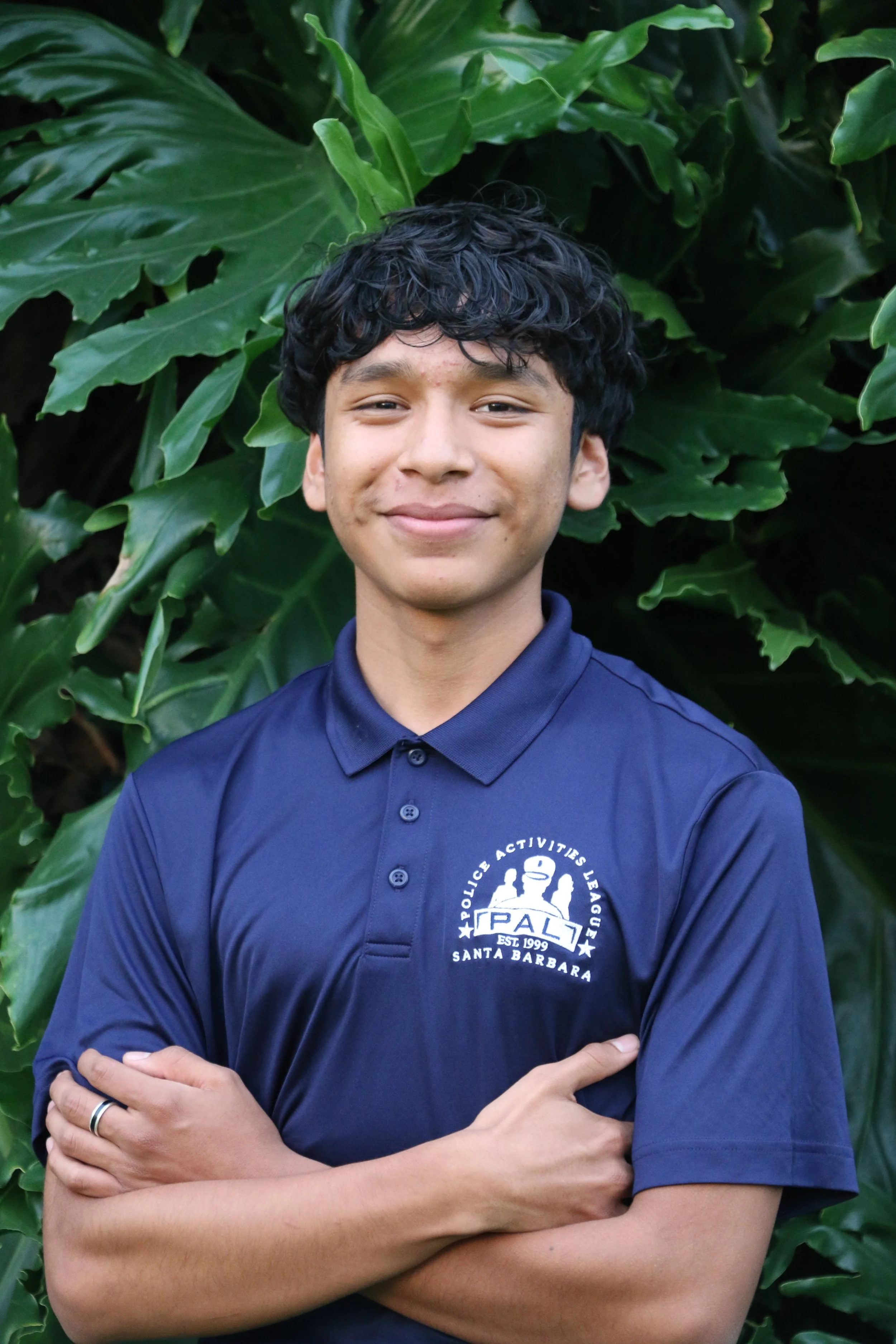 A young boy smiling outdoors, wearing a navy blue polo shirt with a PAL Santa Barbara logo, a silver chain necklace, and standing in front of green foliage and a tree.