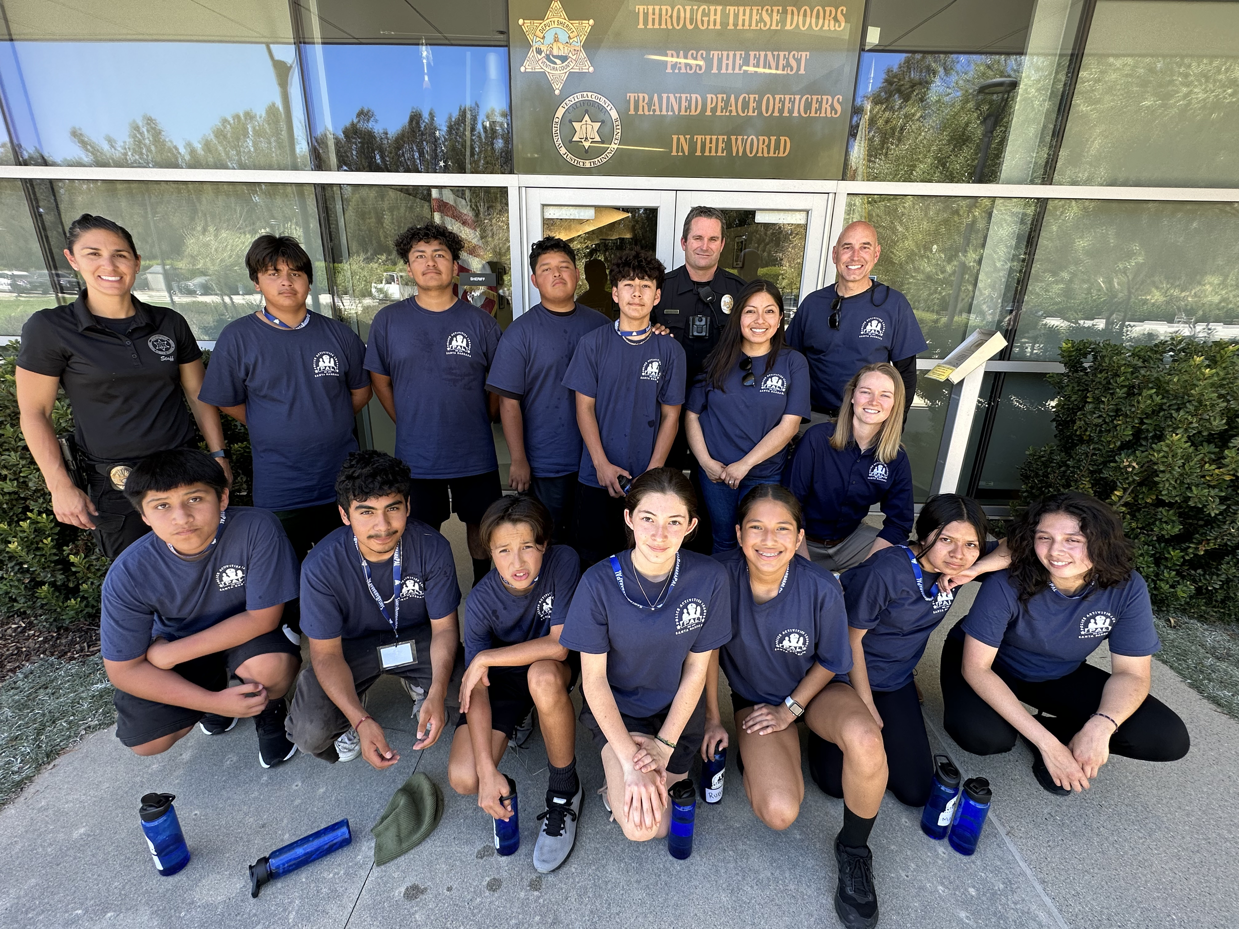 Group of young skateboarding students and police officers standing outside in front of a building with a sign that reads, 'Through these doors pass the finest trained peace officers in the world.' The students are wearing matching navy t-shirts with a logo, some are kneeling in the front row, and some are standing behind. Four water bottles are on the ground. The setting appears to be sunny with trees reflected in the glass.