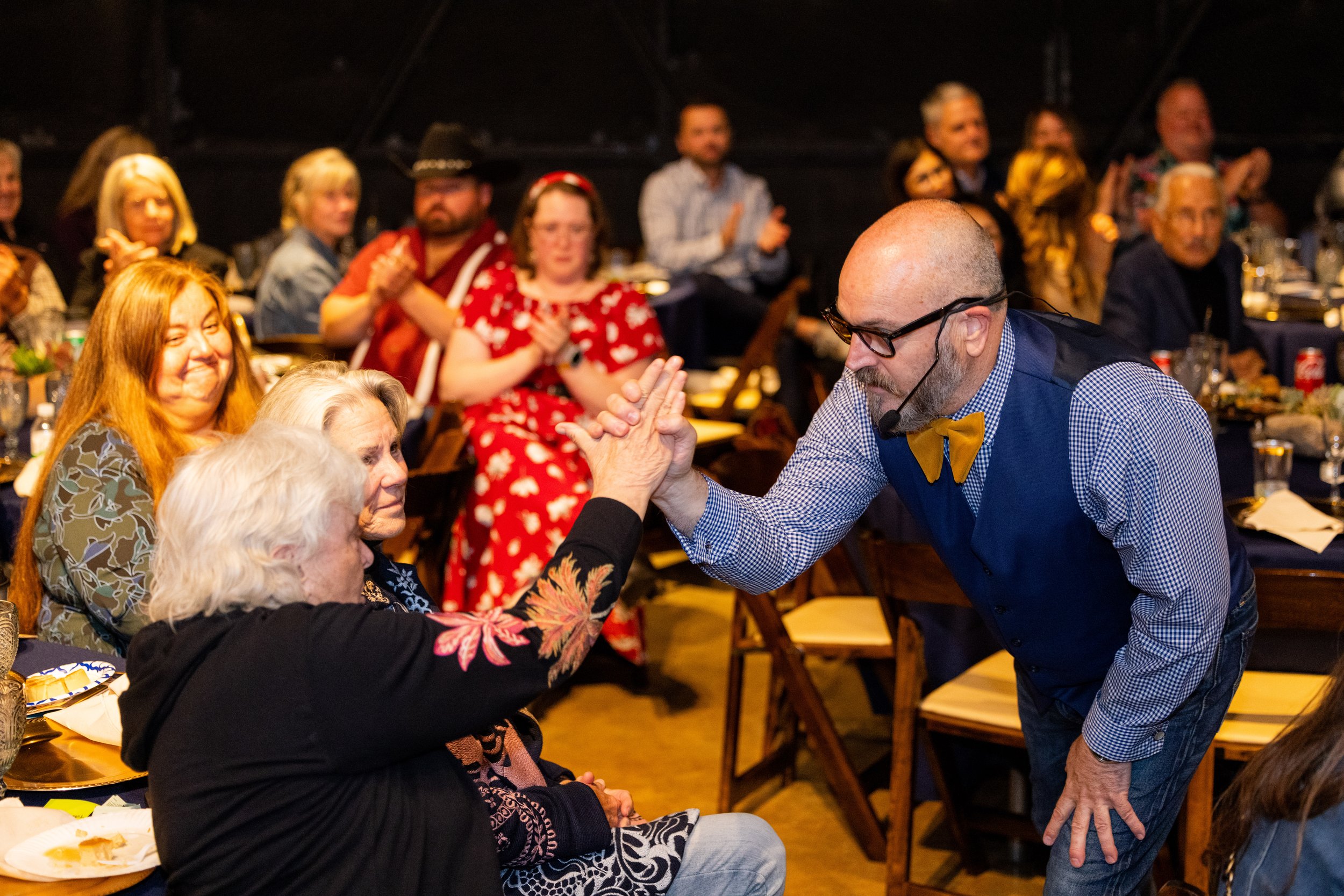 A man with glasses and a beard, wearing a blue vest and a yellow bow tie, is high-fiving an elderly woman with white hair, sitting at a dinner event. The background shows other people clapping and smiling at the gathering.
