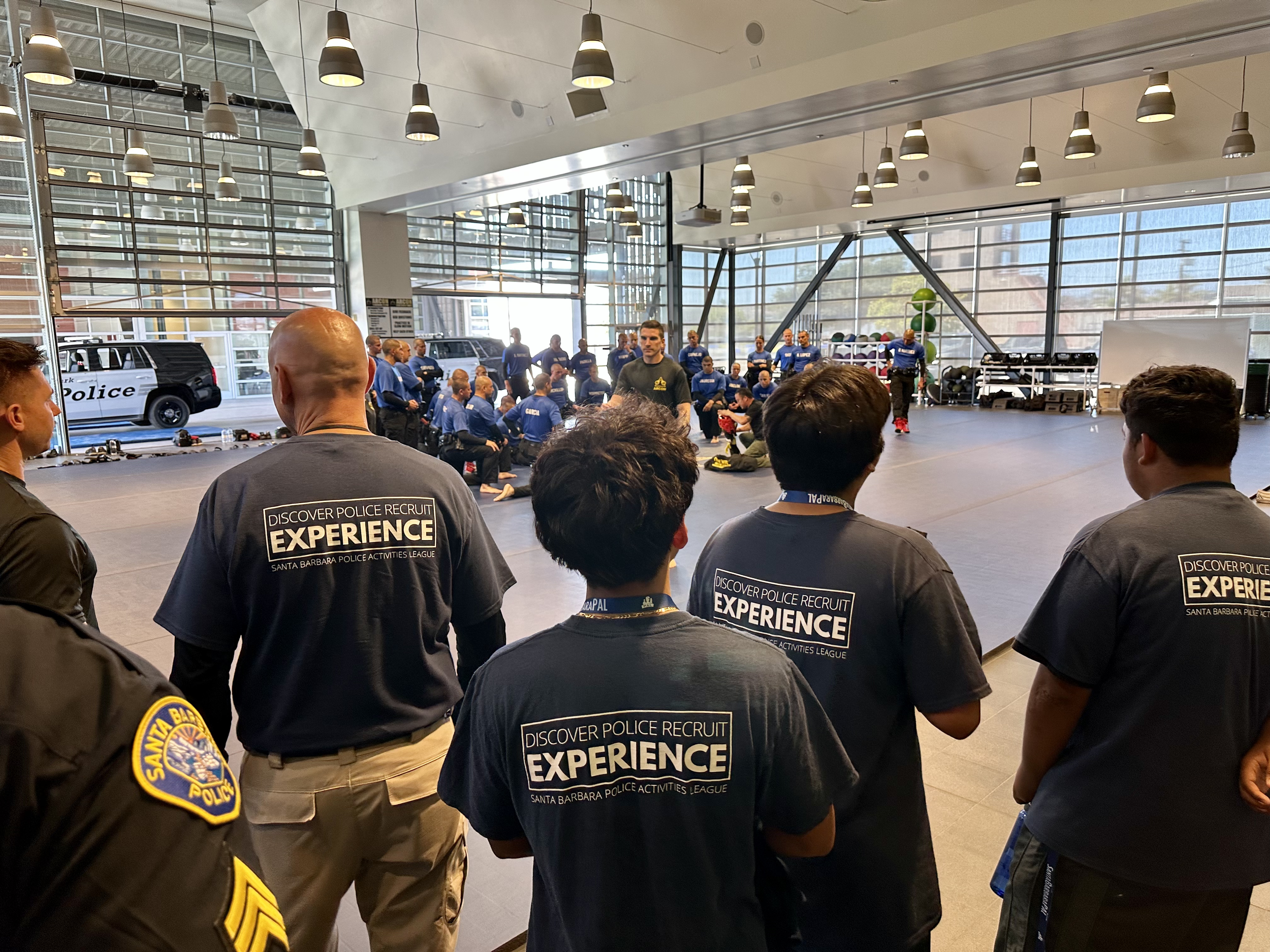Group of police officers in a large indoor training or classroom space, sitting or standing and facing a presentation or demonstration area. The room has large windows, high ceilings with hanging lights, and various gym equipment in the background. Some officers are wearing black shirts with a logo, while others are in police uniforms.