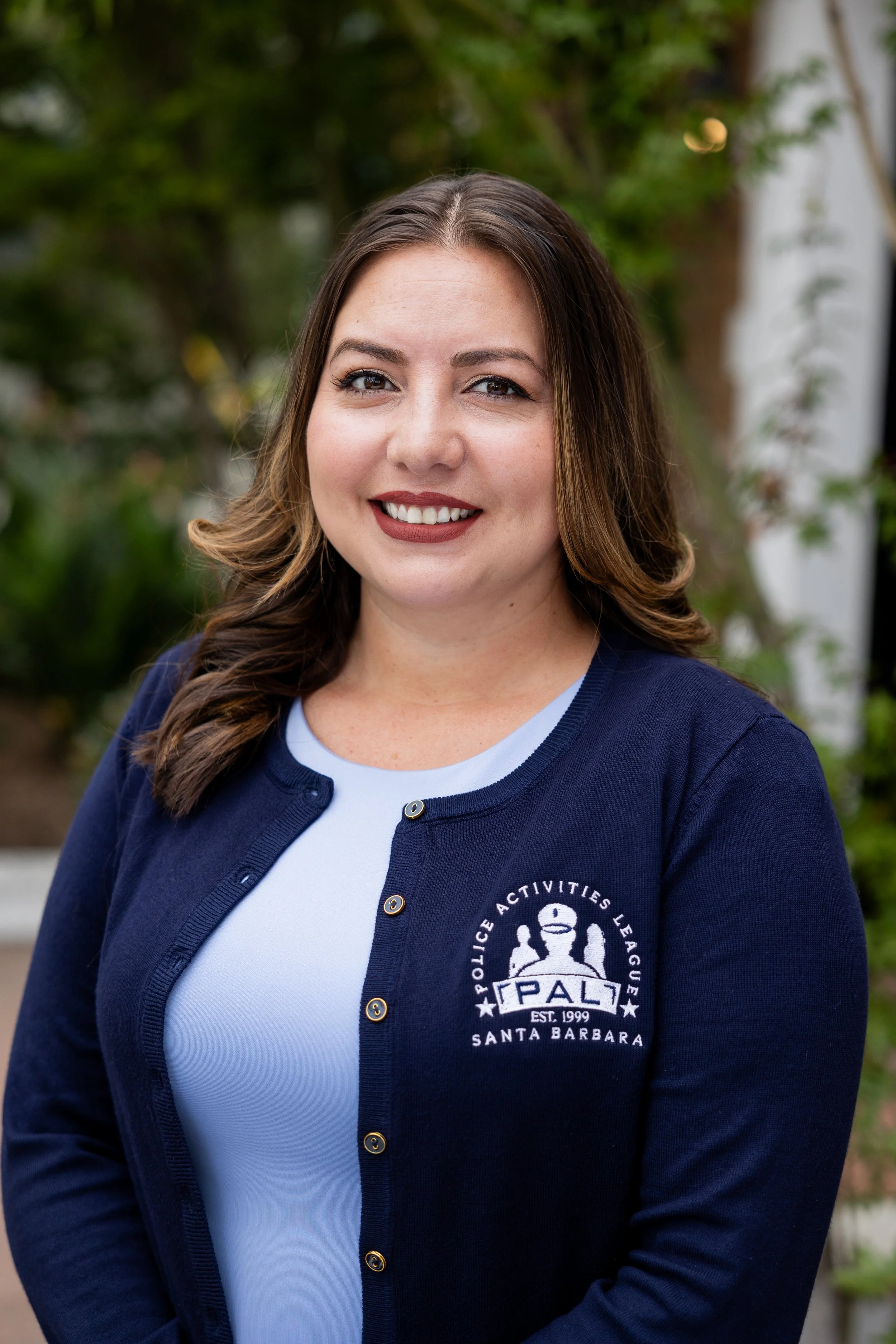 A woman with shoulder-length brown hair, wearing a navy blue cardigan with the Police Activities League logo, standing outdoors in front of greenery, smiling at the camera.
