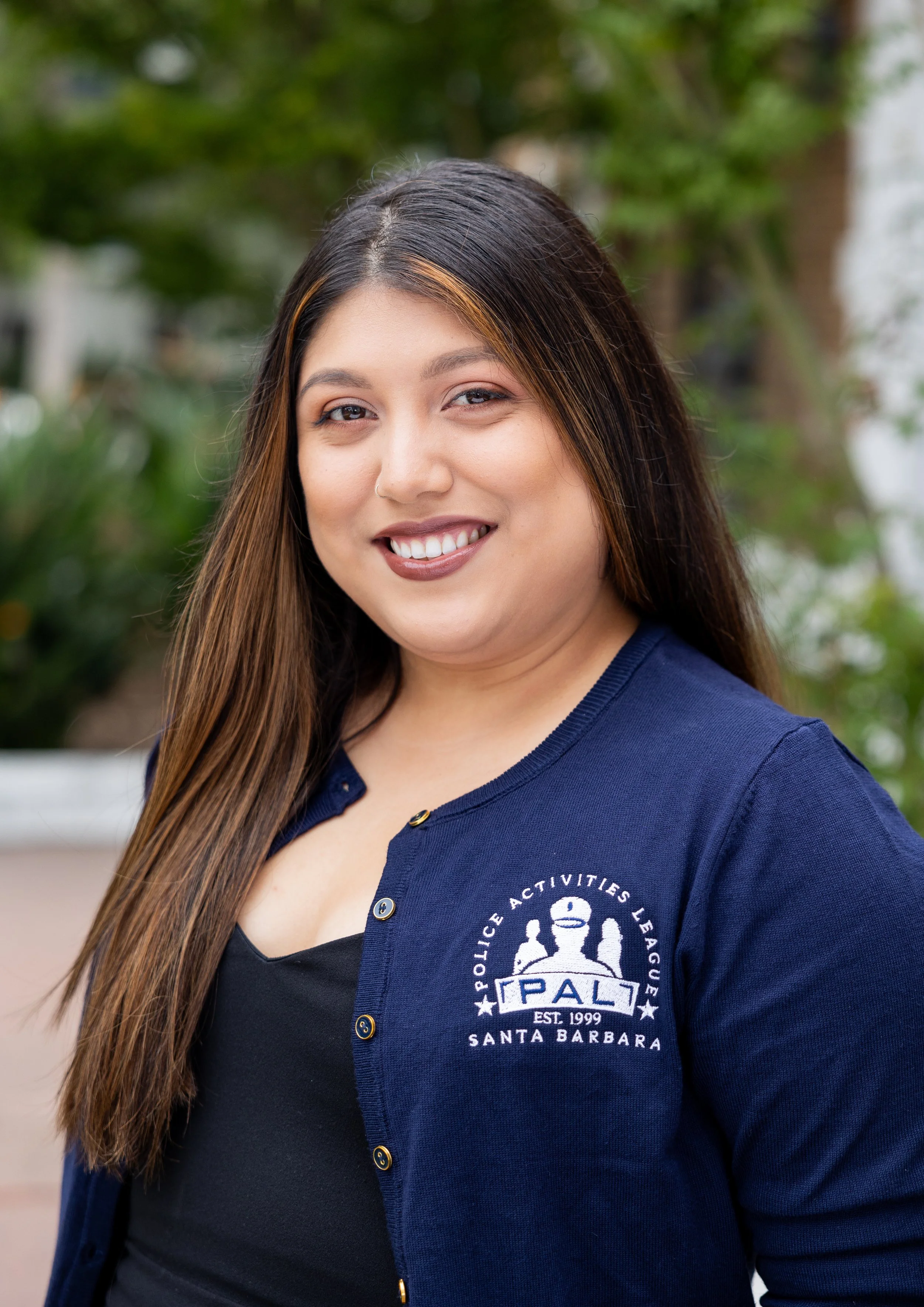 A woman with long brown hair and a warm smile, wearing a blue jacket with the Police Activities League logo, standing outdoors with greenery in the background.